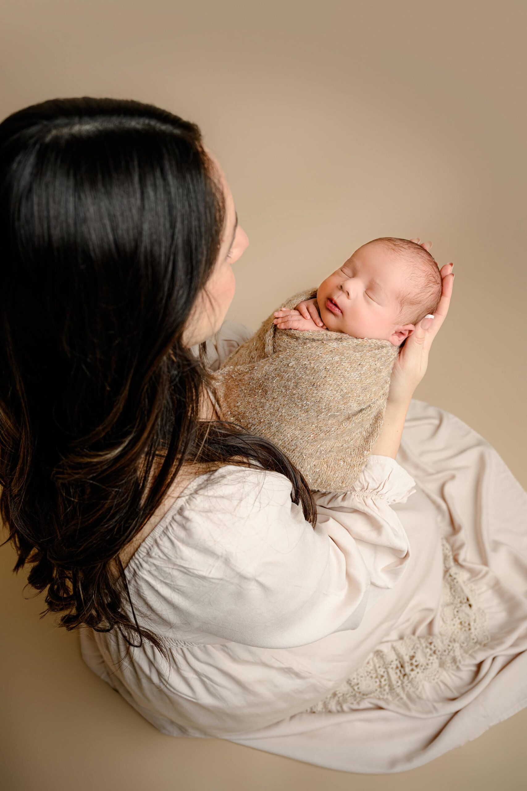 A shot taken over mom's shoulder, capturing the moment she lovingly gazes down at her newborn son, who is peacefully sleeping in her arms. The soft brown background adds to the warmth and tenderness of the newborn session scene.
