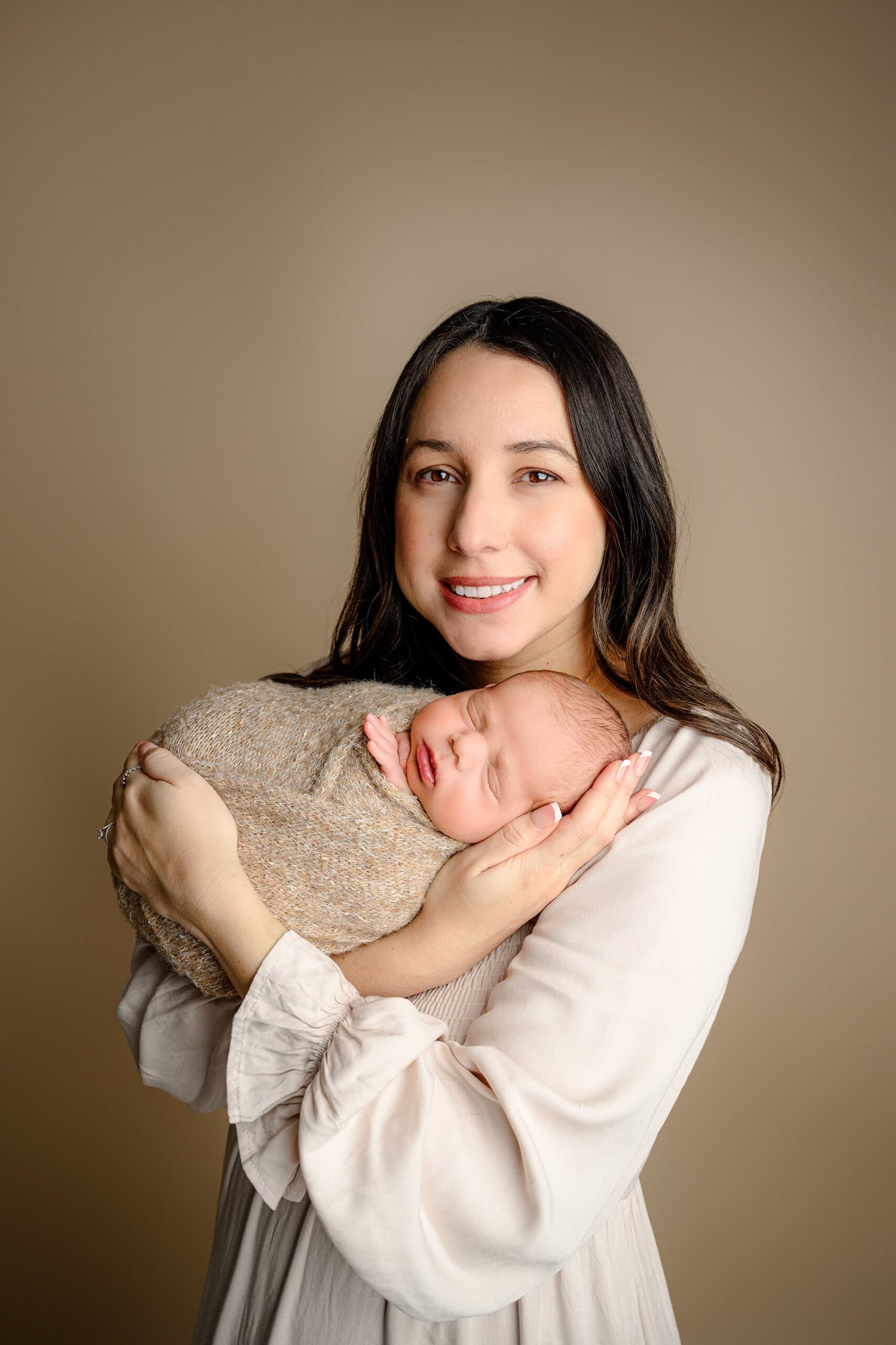 Monroe, NC Mom, dressed in a light brown dress, cradles her newborn baby boy in her arms, looking toward the camera. The baby, wrapped in a brown wrap, is nestled close to her shoulder, showcasing their bond in a peaceful, intimate moment.