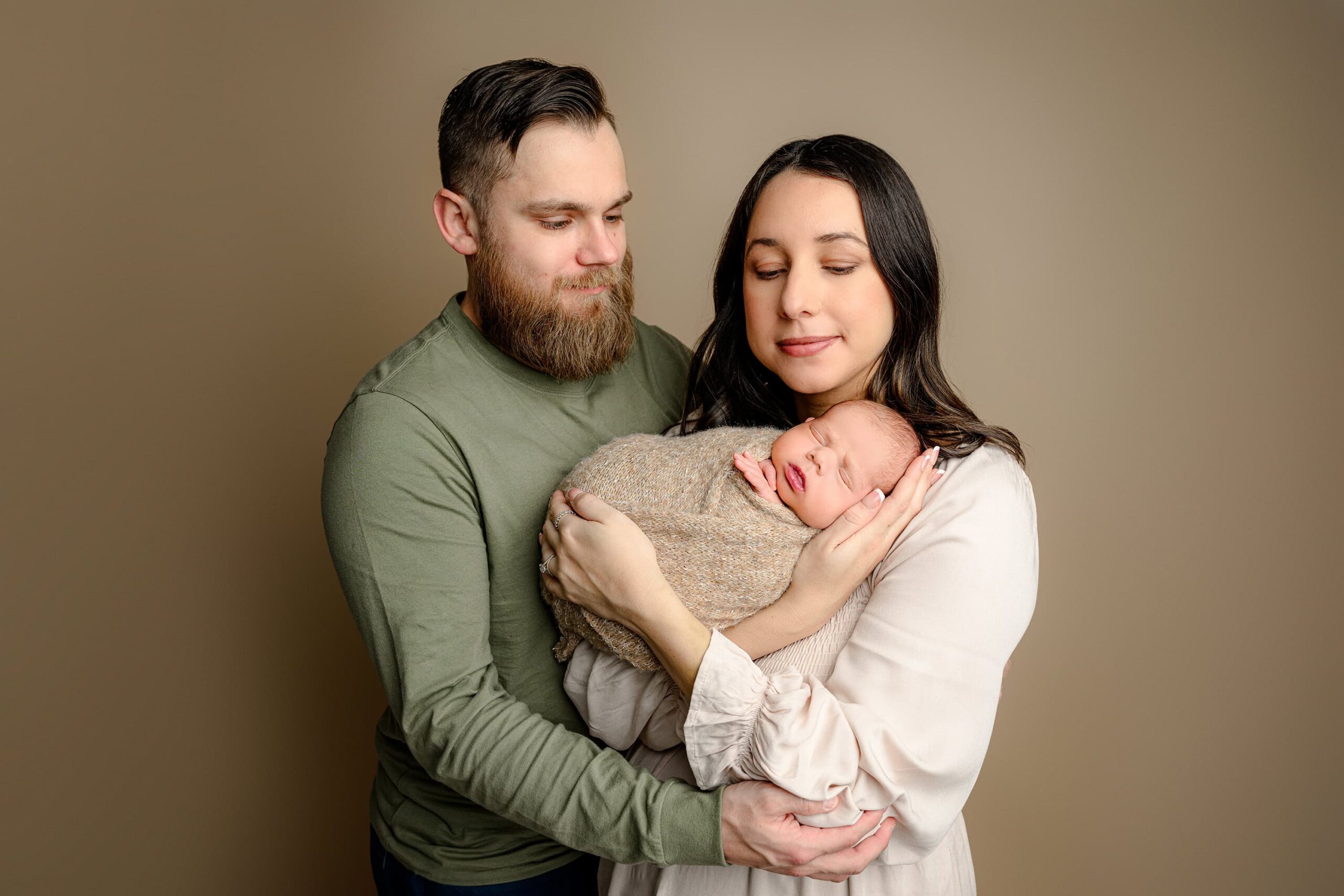 Monroe, NC family portrait featuring a mom in a light brown dress with long dark hair, cradling her newborn baby wrapped in a brown wrap. Dad, wearing green, stands beside her, and they both look down lovingly at their baby. The background is a warm brown, creating a cozy and intimate family moment.