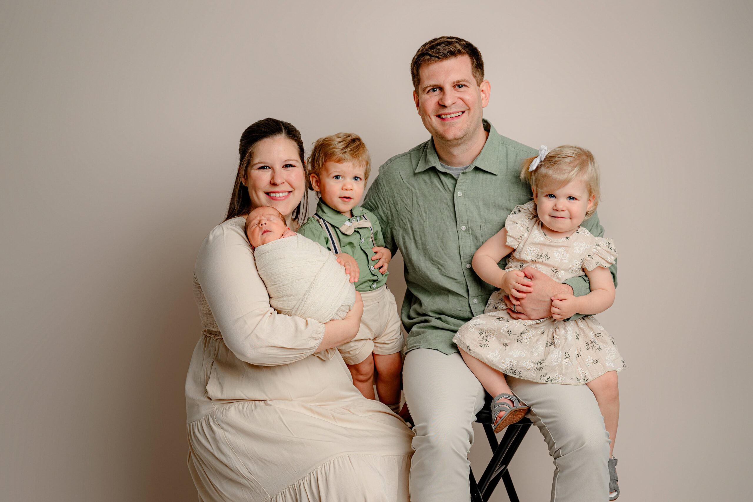 West Charlotte area parents at my studio, with mom in cream holding a newborn baby boy wrapped in cream, twin toddlers sitting with dad—boy in a green shirt, suspenders, and khaki shorts, and girl in a tan floral dress with a white bow in her blonde hair.