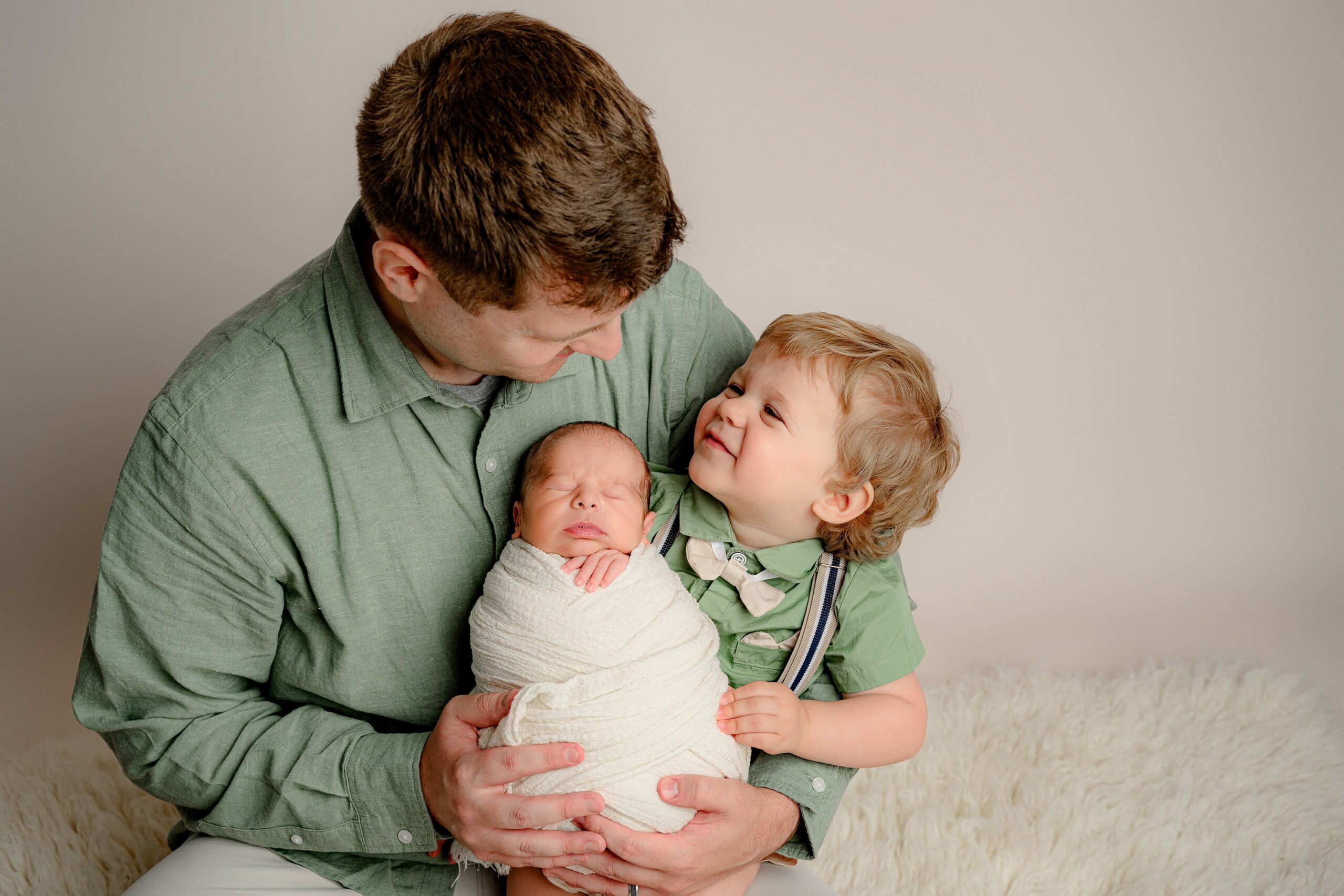 Charlotte, NC area Father and son in matching green shirts hold a newborn wrapped in a cream blanket, sharing a tender moment as they smile at each other.