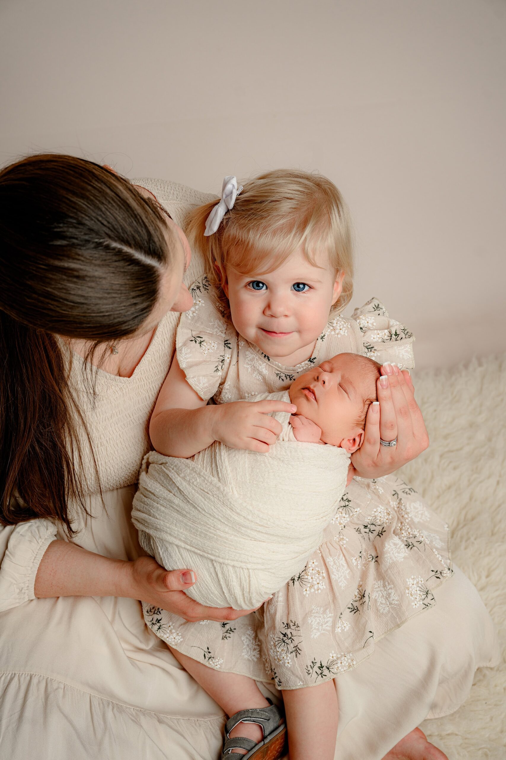Mother in a cream dress holding her newborn baby boy wrapped in cream and her toddler daughter in a tan floral dress with a white bow, as the toddler looks at the camera.