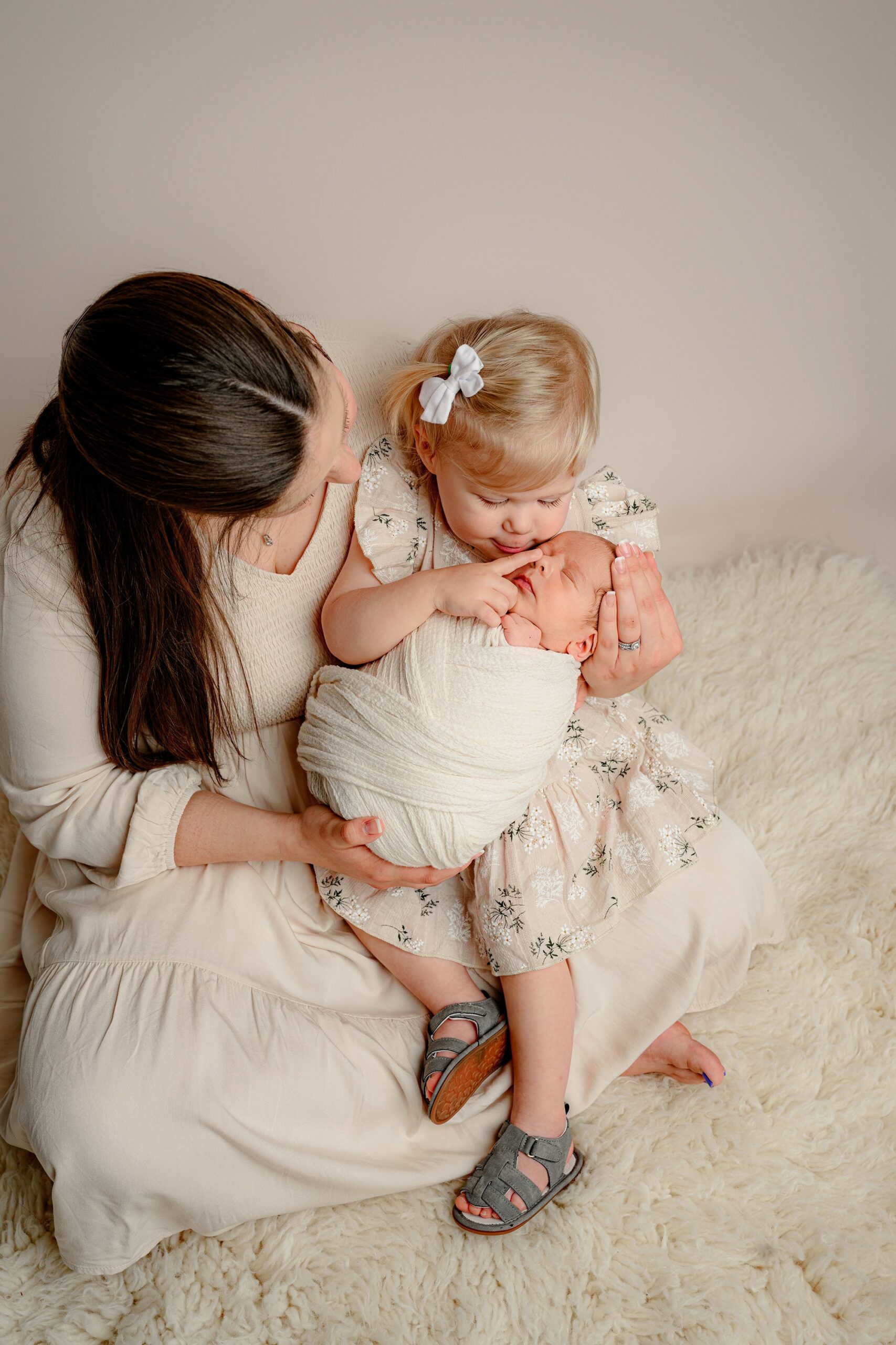 Mother in a cream dress holding her newborn baby boy wrapped in cream and her toddler daughter, who gently touches her baby brother’s nose.