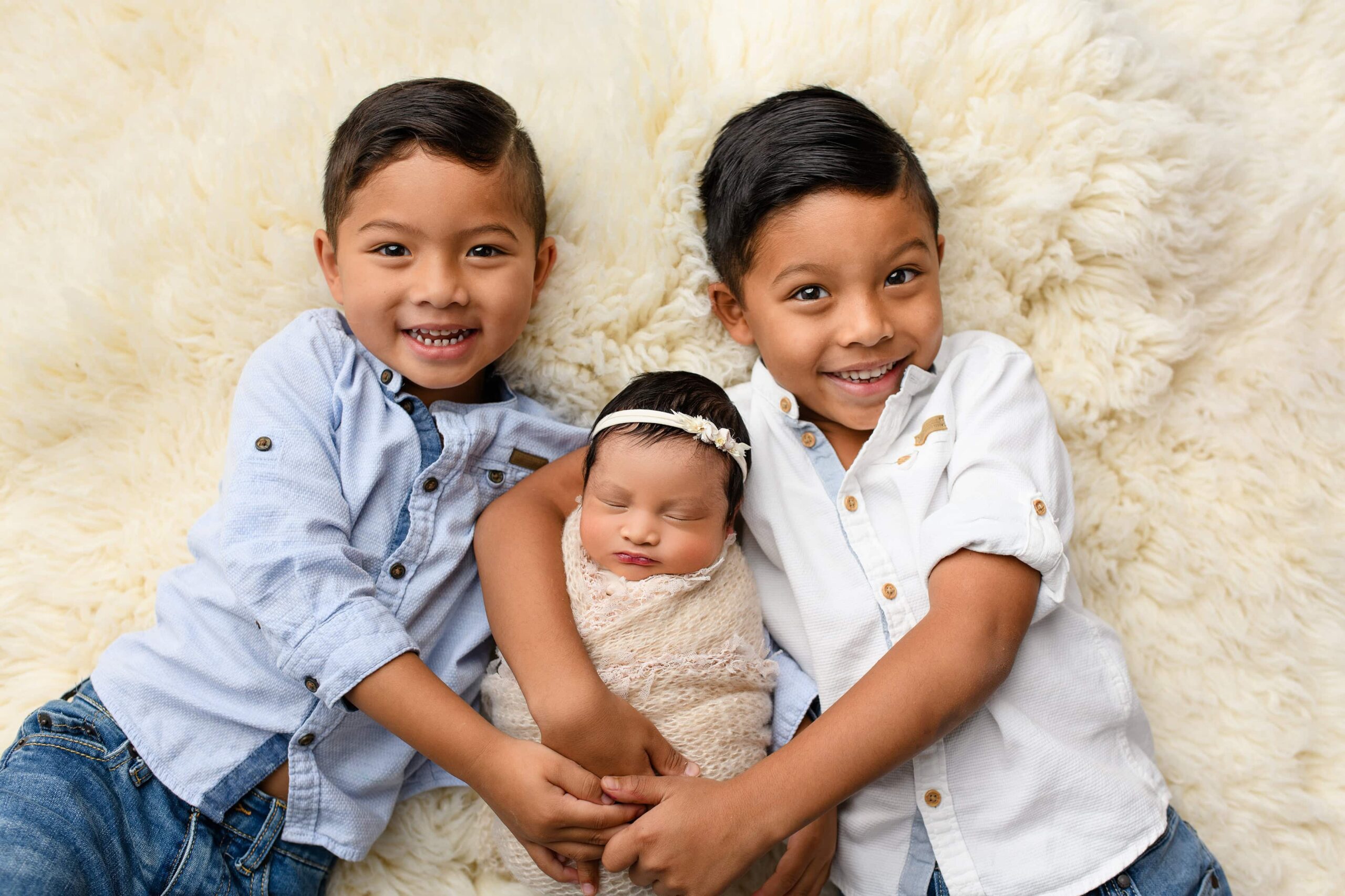 Two brothers, one in a blue shirt and the other in white, hold their newborn sister wrapped in a cream blanket with a matching bow, lying on a soft cream fur layer in the waxhaw studio space.