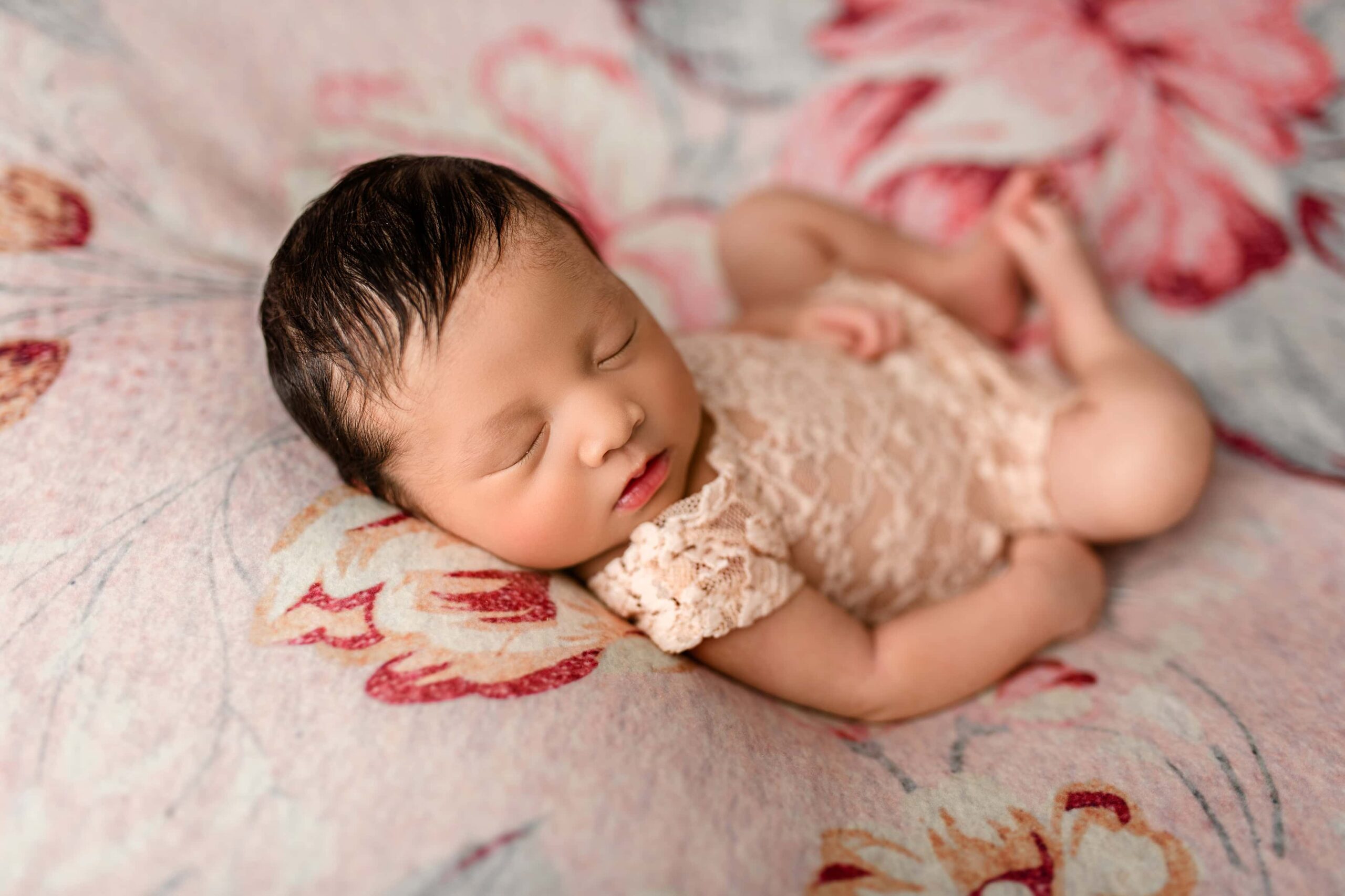 Newborn baby girl in a peach lace outfit, lying on a floral background with teal and pink tones, posed on her back with legs curled.