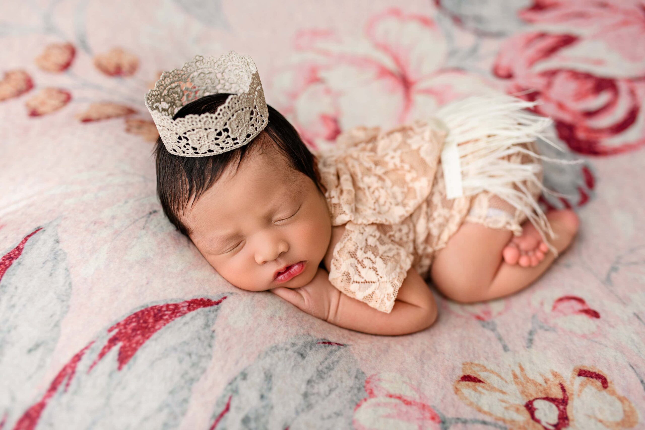Newborn baby girl curled up on her belly, wearing a delicate crown, showcasing her thick dark hair on a teal and pink floral background.