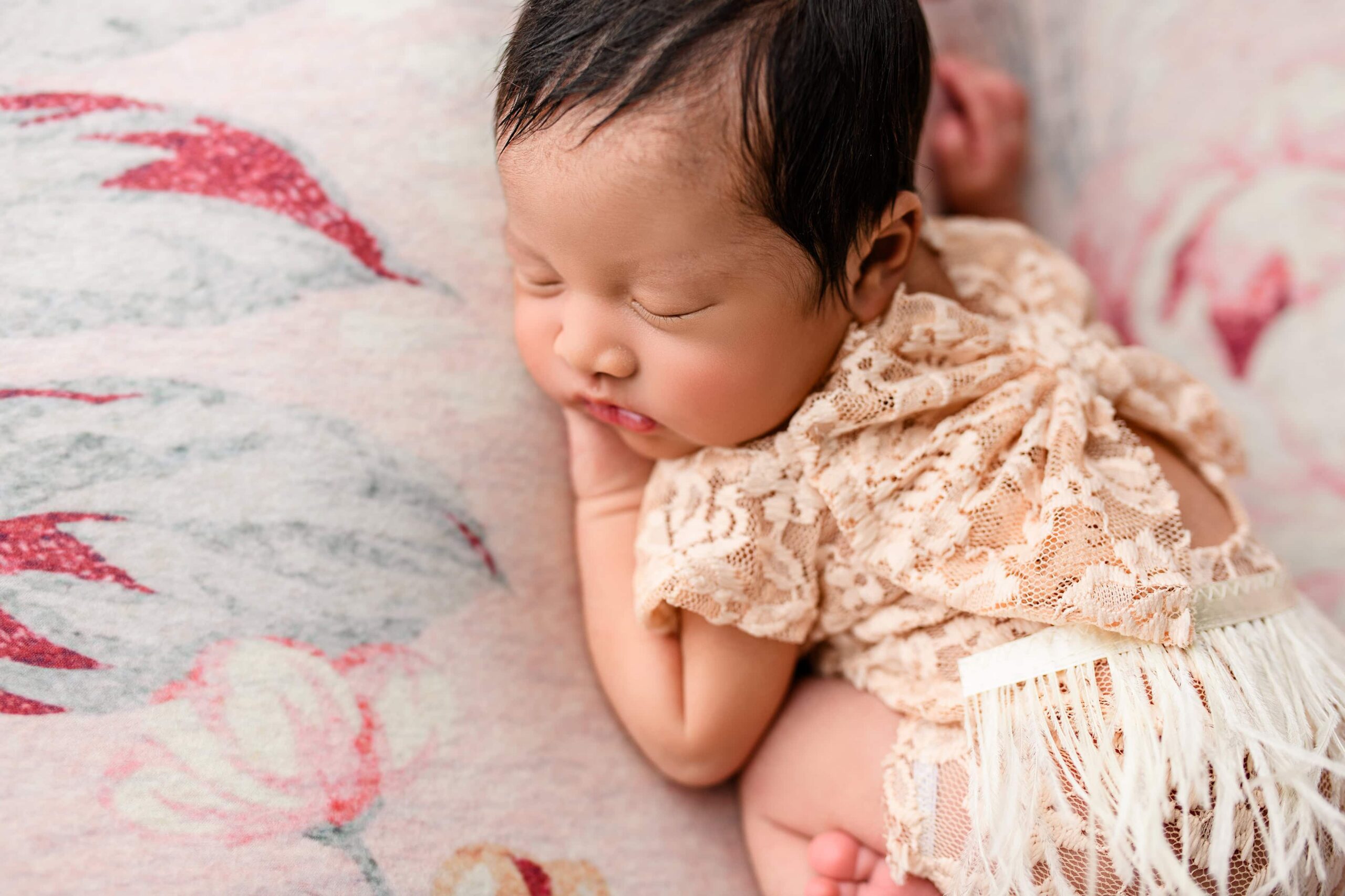Overhead shot of a newborn baby girl on a floral backdrop, highlighting her cheeks and thick dark hair, dressed in peach lace.