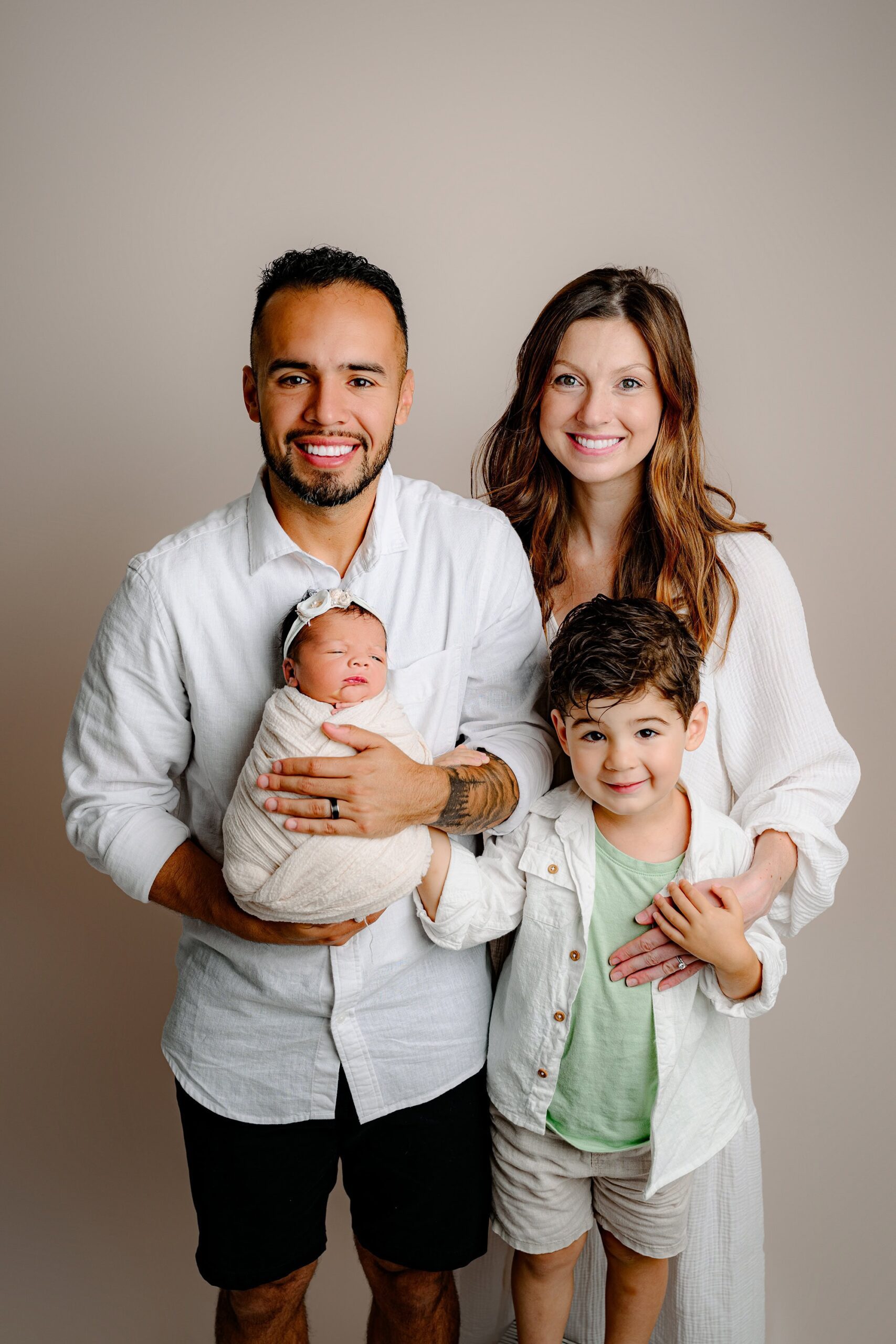 Little boy with his Charlotte area parents dressed in white and cream, as dad holds their newborn daughter wrapped in white with a bow on her dark hair.