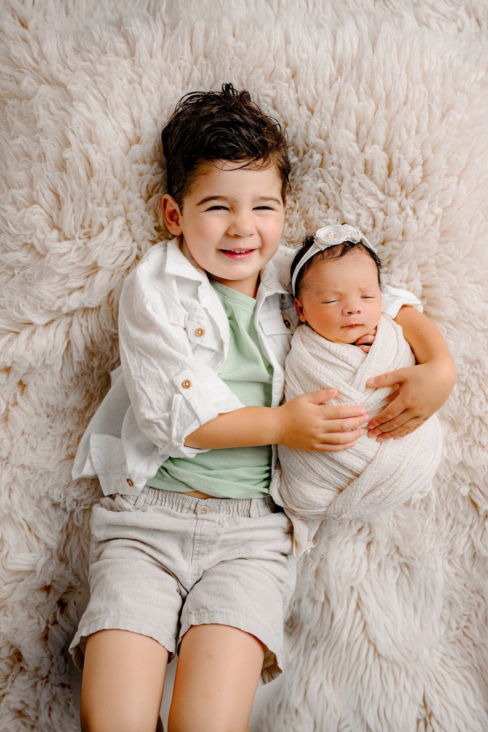 Little brother from a Charlotte, NC family wearing a white button-down over a green shirt and khaki shorts, smiling at the camera while holding his newborn sister wrapped in white on a cream fur.