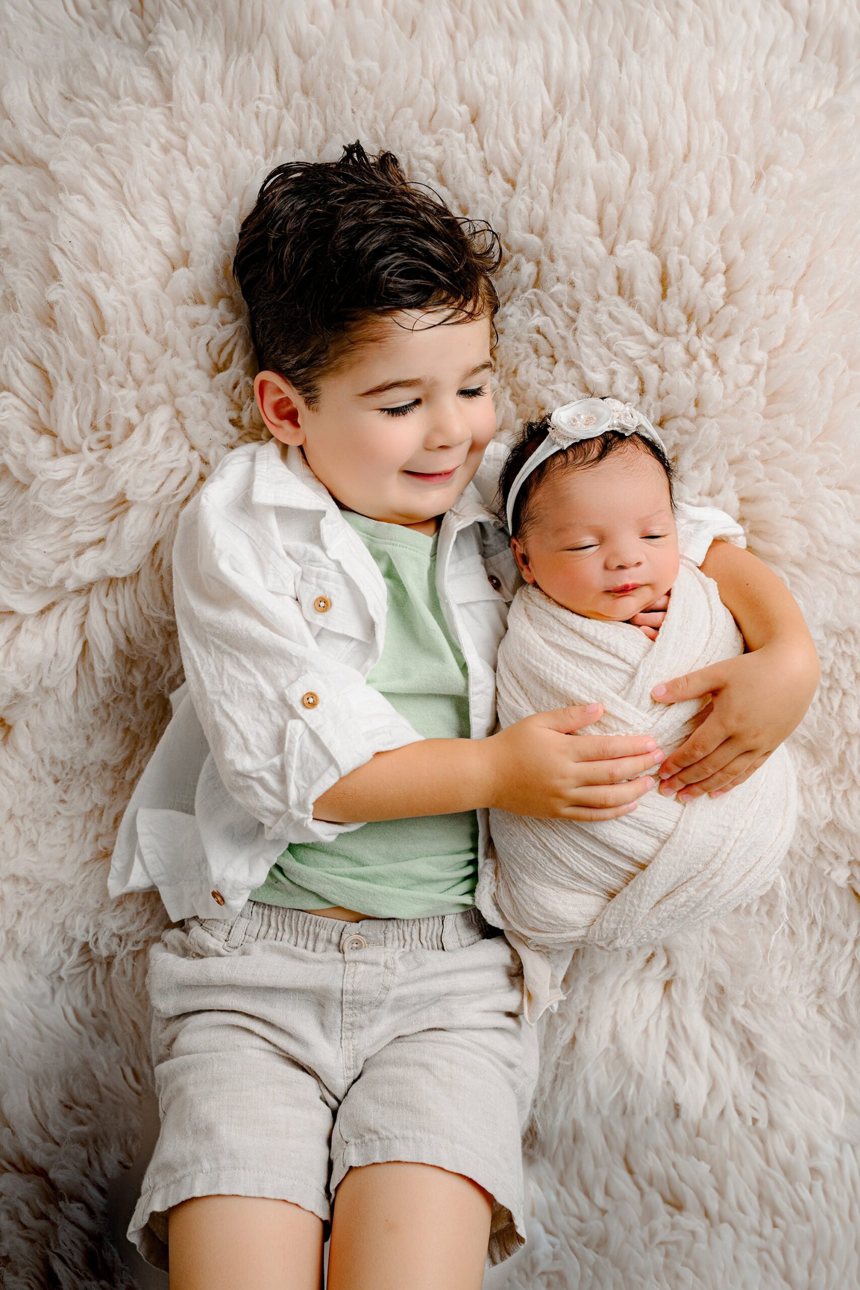 Little brother from a Charlotte, NC family smiling warmly at his newborn sister, who is wrapped in white and lying on a cream fur layer.
