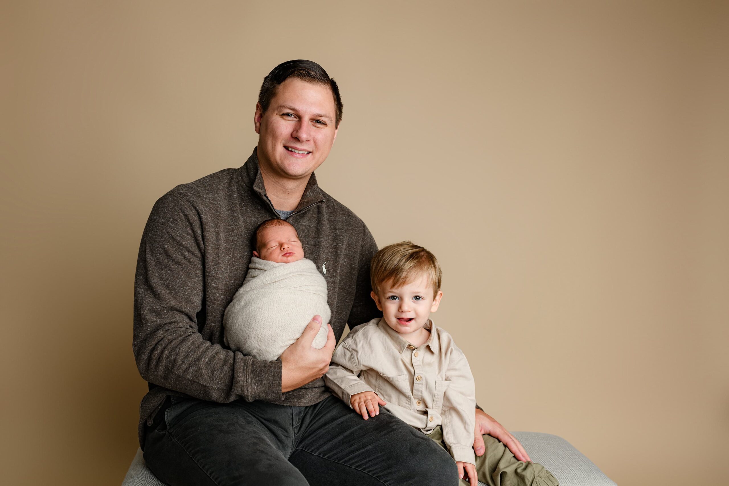 Lancaster, SC dad in a brown shirt holding his newborn baby boy wrapped in tan, with his toddler son in khaki and green sitting beside him, both posed in a Waxhaw studio.