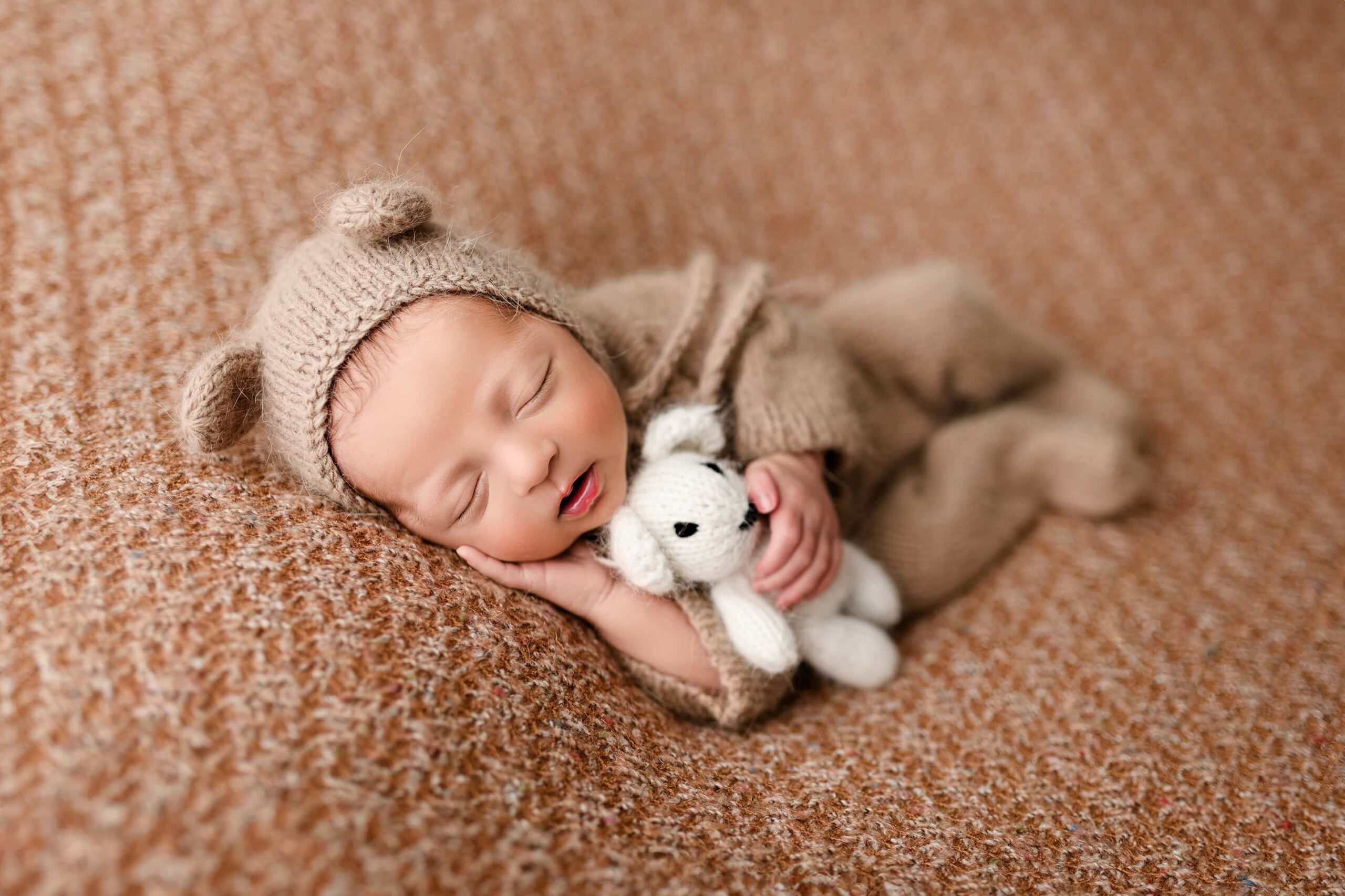 Newborn baby wearing a tan outfit and a bear bonnet with ears, posed on a brownish-orange fabric, lying on his side while holding a teddy bear with one hand and resting the other under his cheek.