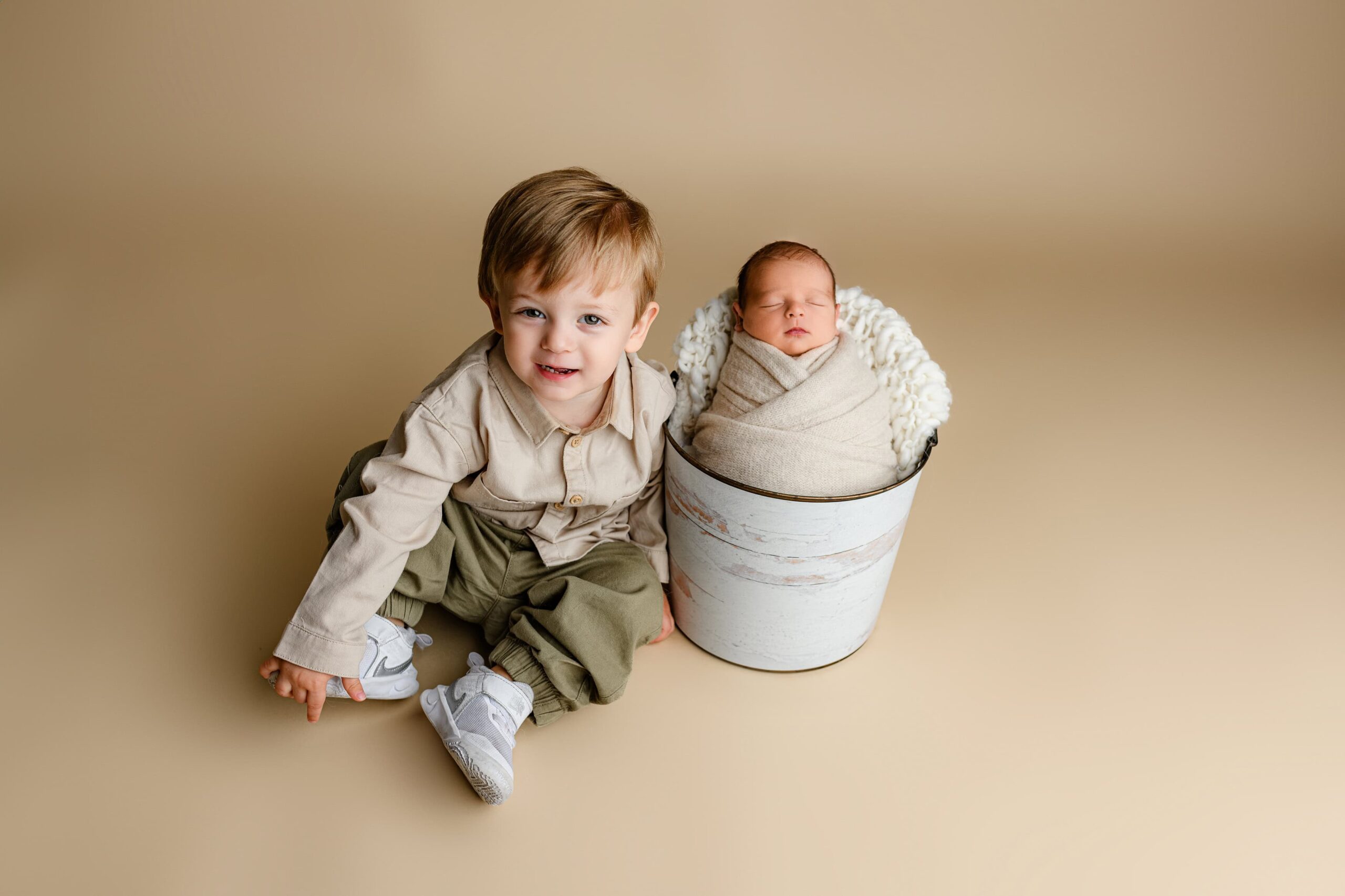 Toddler boy from Waxhaw sitting next to his newborn brother wrapped in cream in a bucket, both looking at the camera on a cream layer.