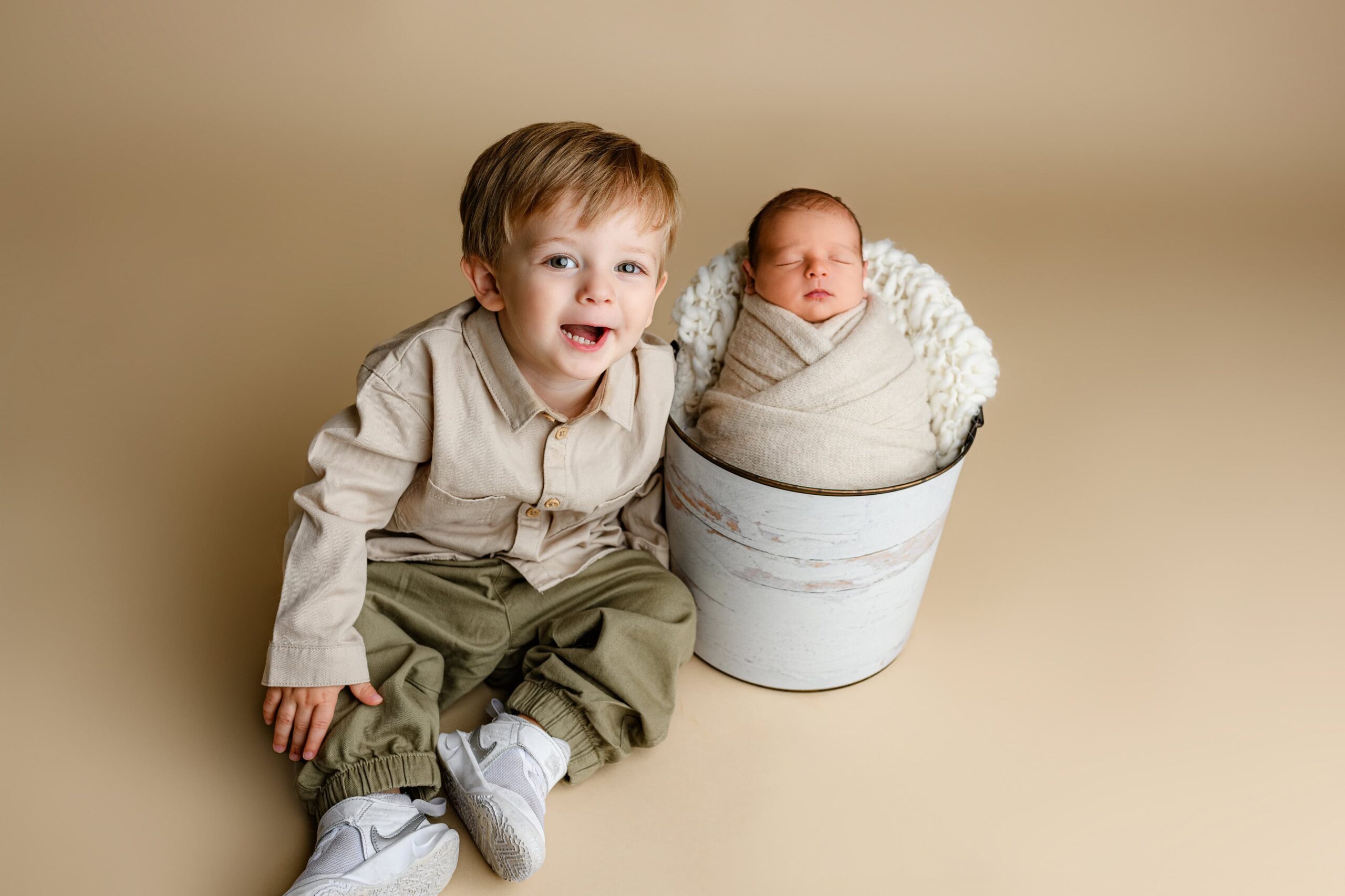 Toddler boy from Waxhaw in a khaki shirt and green pants smiling and laughing beside his newborn baby brother wrapped in cream, posed in a bucket.