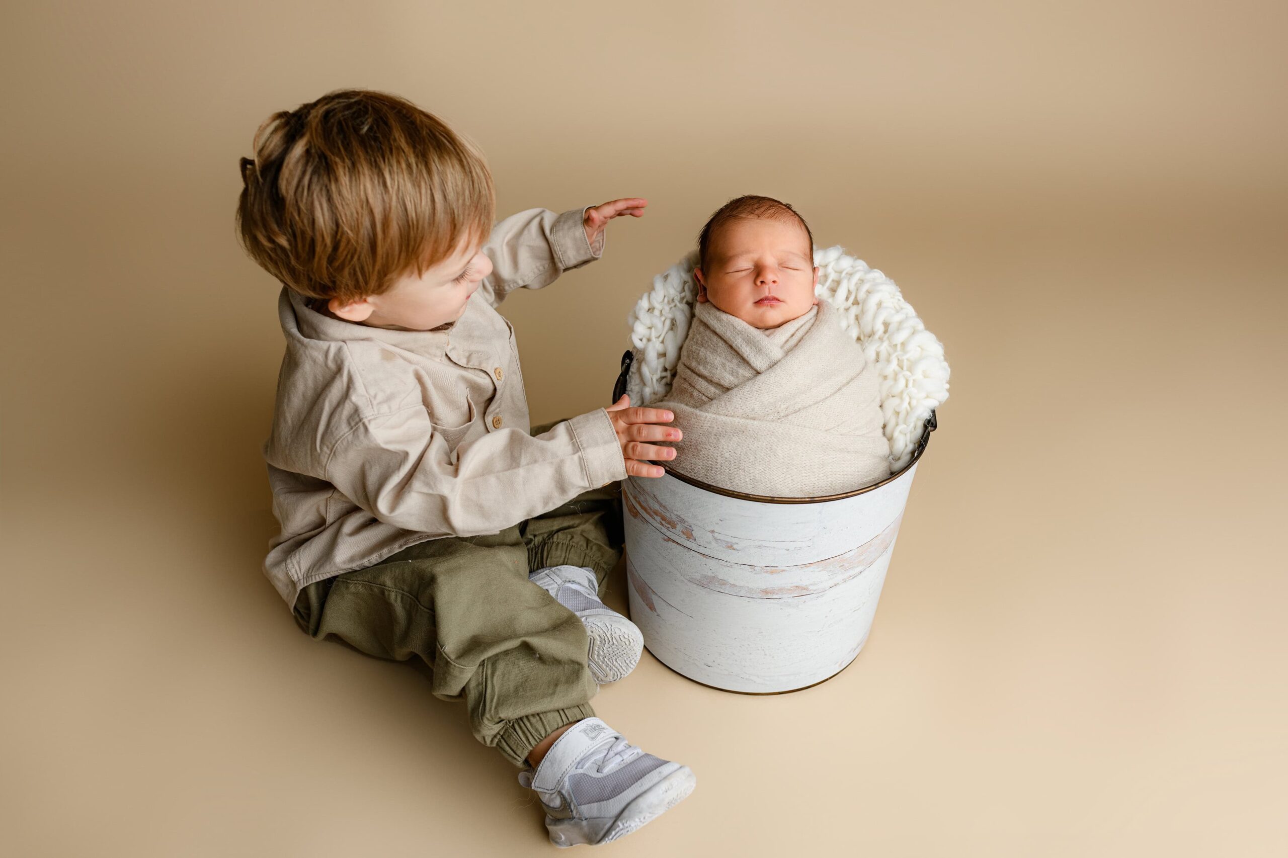 Toddler boy from Waxhaw in khaki and green reaching out to hug his newborn baby brother, who is wrapped in cream and nestled in a bucket.