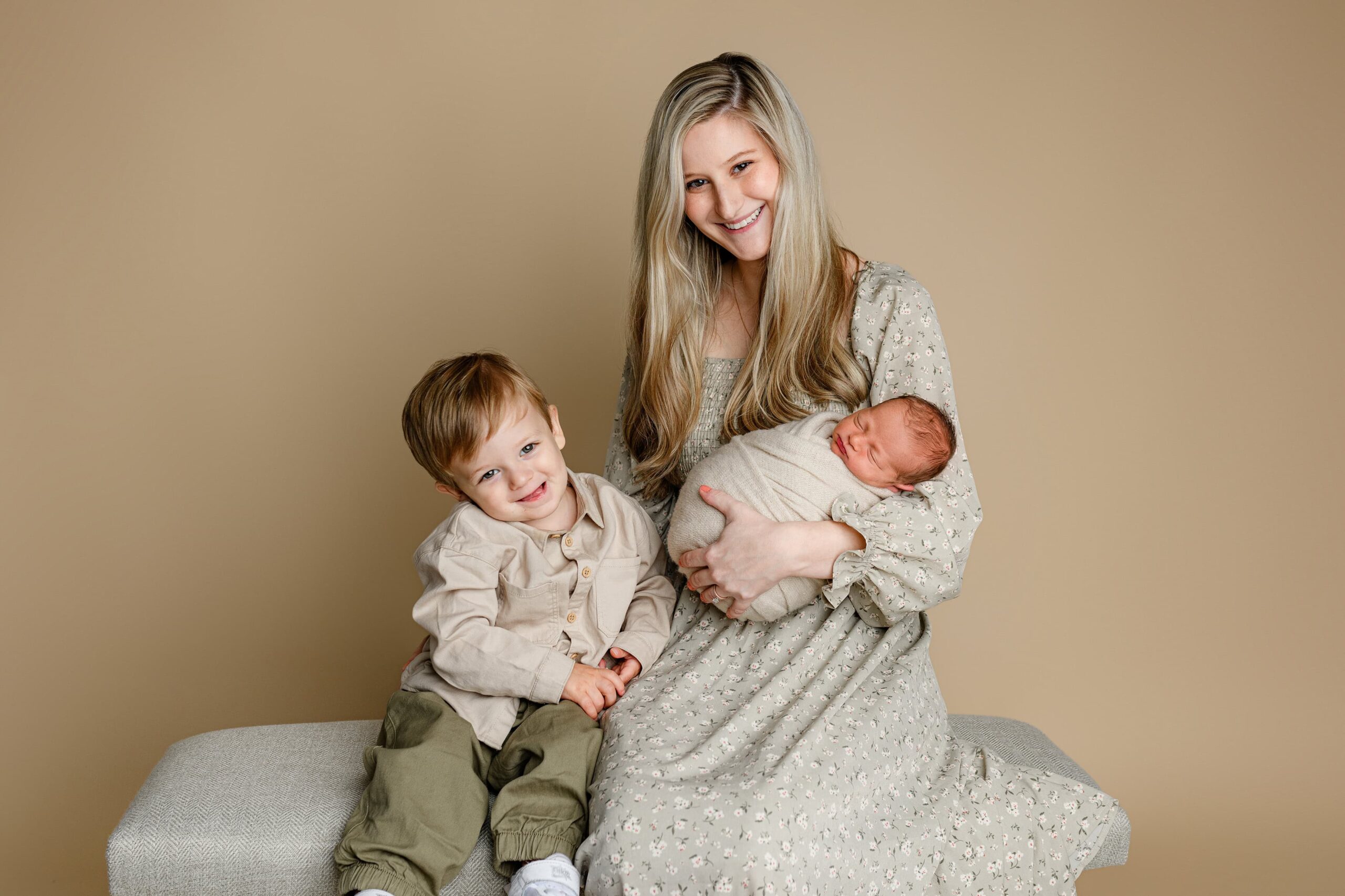 Lancaster, SC mom in a cream dress smiling at the camera while holding her newborn baby boy wrapped in tan, as her toddler son grins brightly at the photographer.