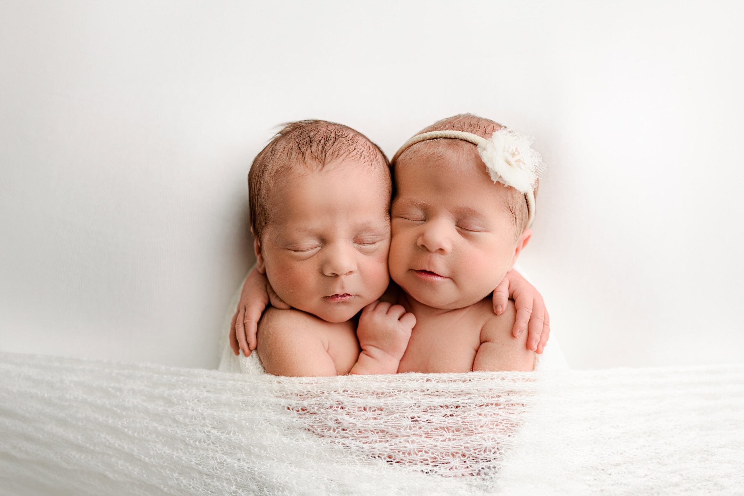 Charlotte area newborn boy and girl twins cuddled together on a white background, wrapped in a delicate white wrap. The girl wears a white flower headband while the boy is without headgear.