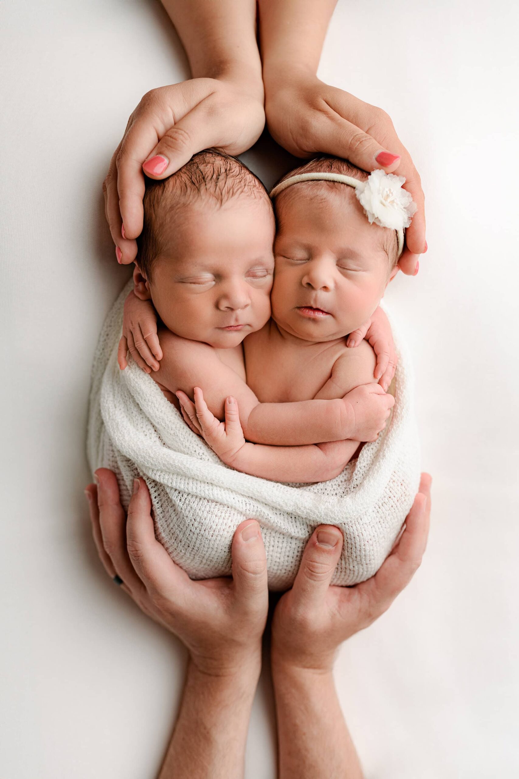 Charlotte area family newborn boy and girl twins resting on a white background, wrapped in a white wrap, with parents' hands included. Mom's hands gently rest over the twins' heads, while Dad's hands hold the bottom of the wrap.