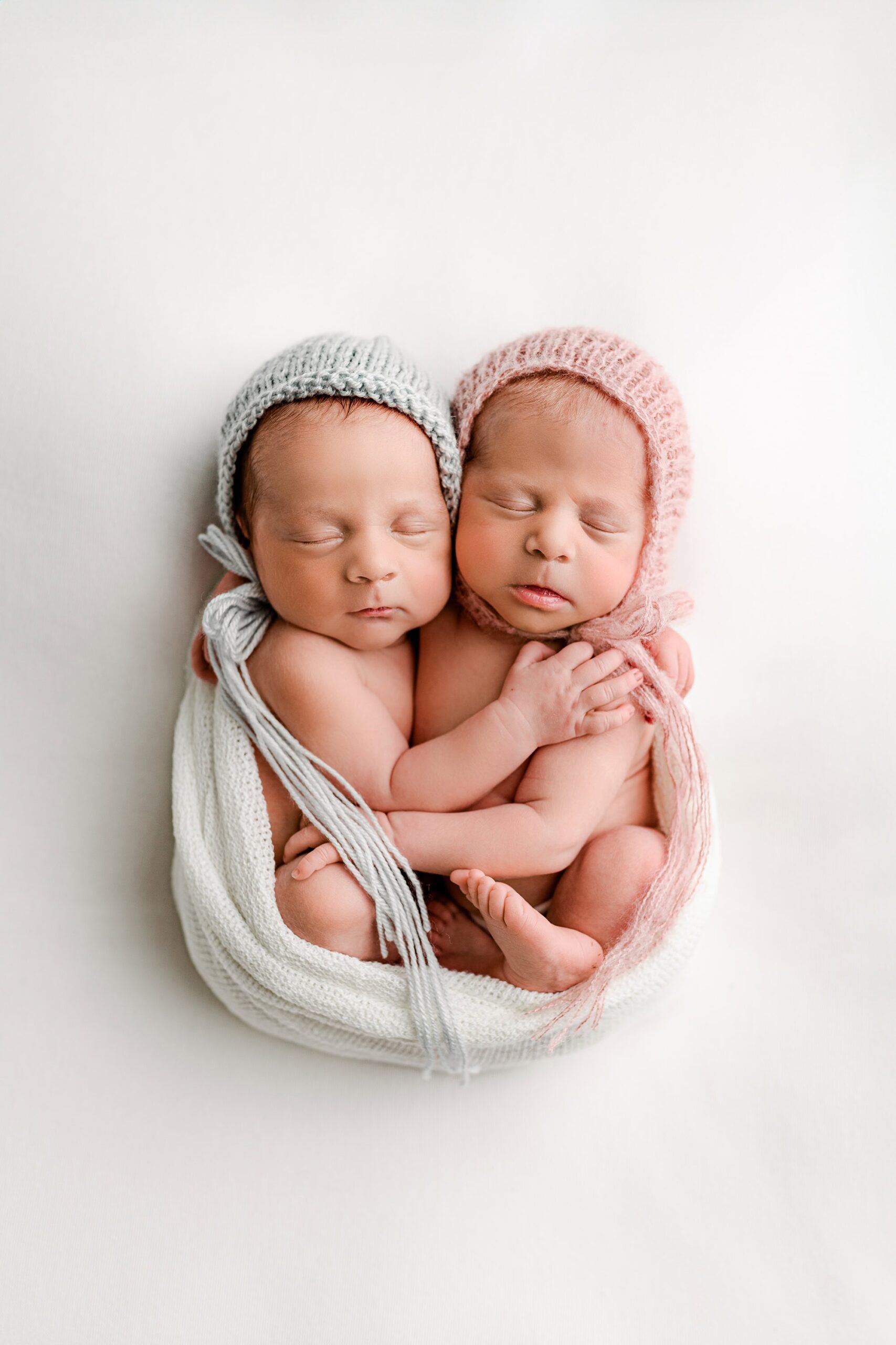 Newborn boy and girl twins from the Charlotte, NC area cuddled together on a white background, wrapped in a delicate white wrap arranged in an egg-like shape. The girl wears a pink knit bonnet, and the boy wears a blue bonnet.