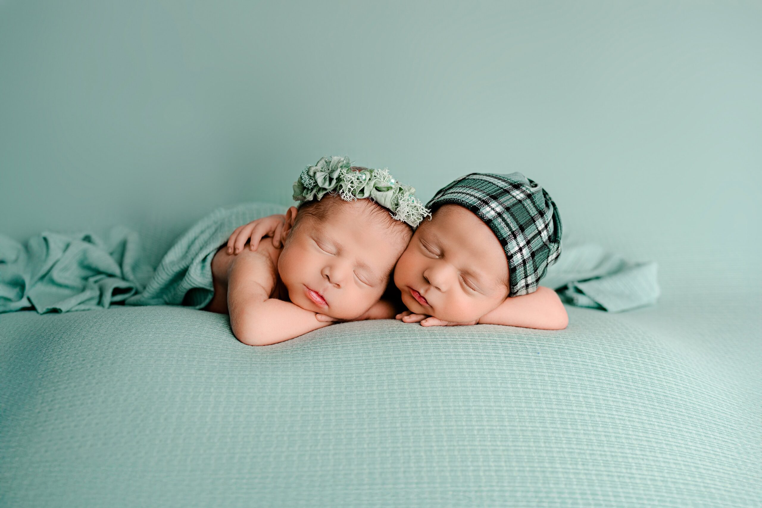 Newborn boy and girl twins from the Charlotte, NC area posed together in a Waxhaw studio on a minty teal background. The girl rests both hands under her left chin, wearing a matching flower and pearls headband, while the boy has his right arm around his sister and left hand under his cheek, wearing a sleepy cap in green and mint tones.