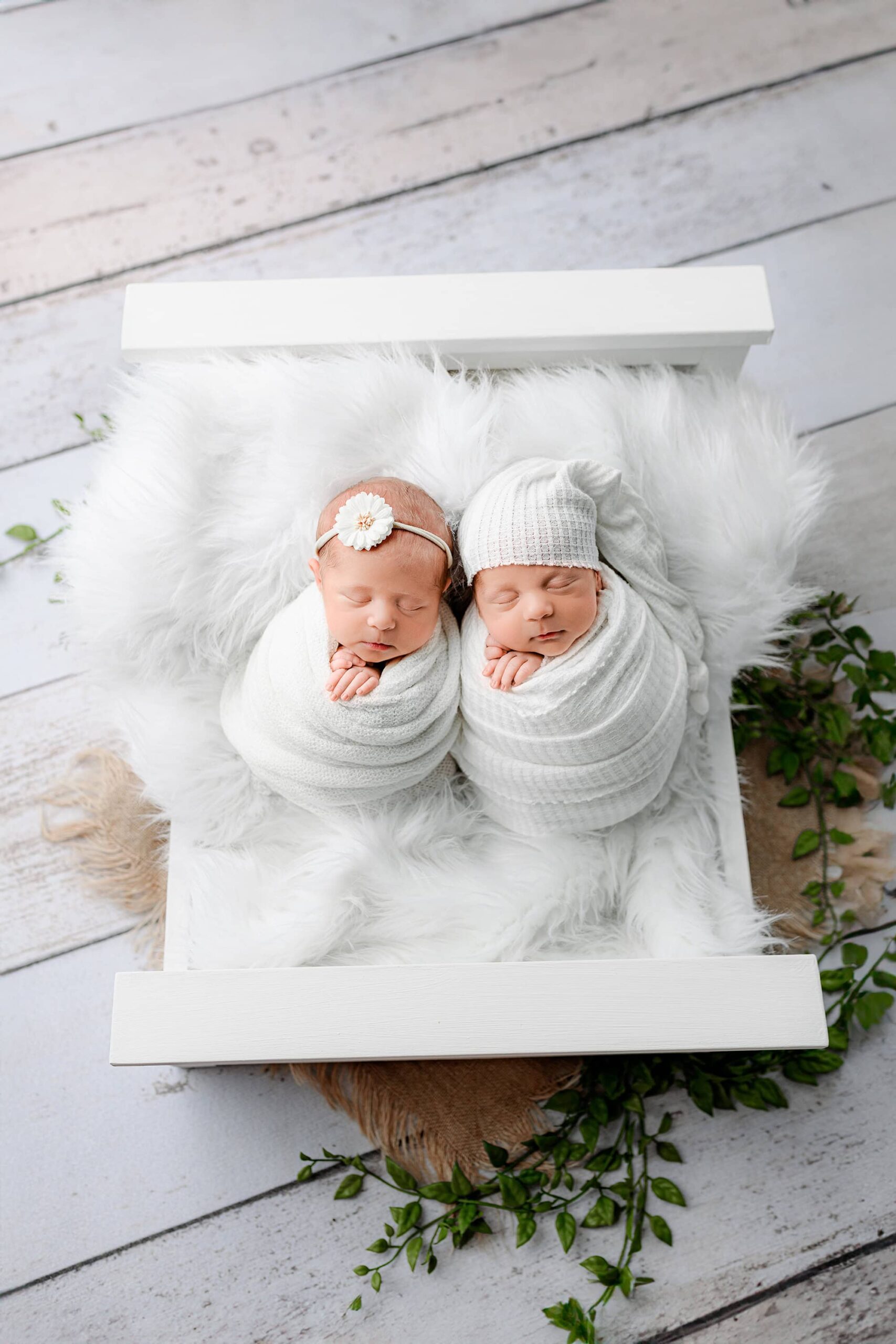 Newborn boy and girl twins wrapped individually in white wraps, posed on a bed with a pulled-back view showing the full bed and natural flooring. The boy is wrapped in a textured wrap with a matching sleepy hat, while the girl is wrapped in a lighter knit with a flower headband.