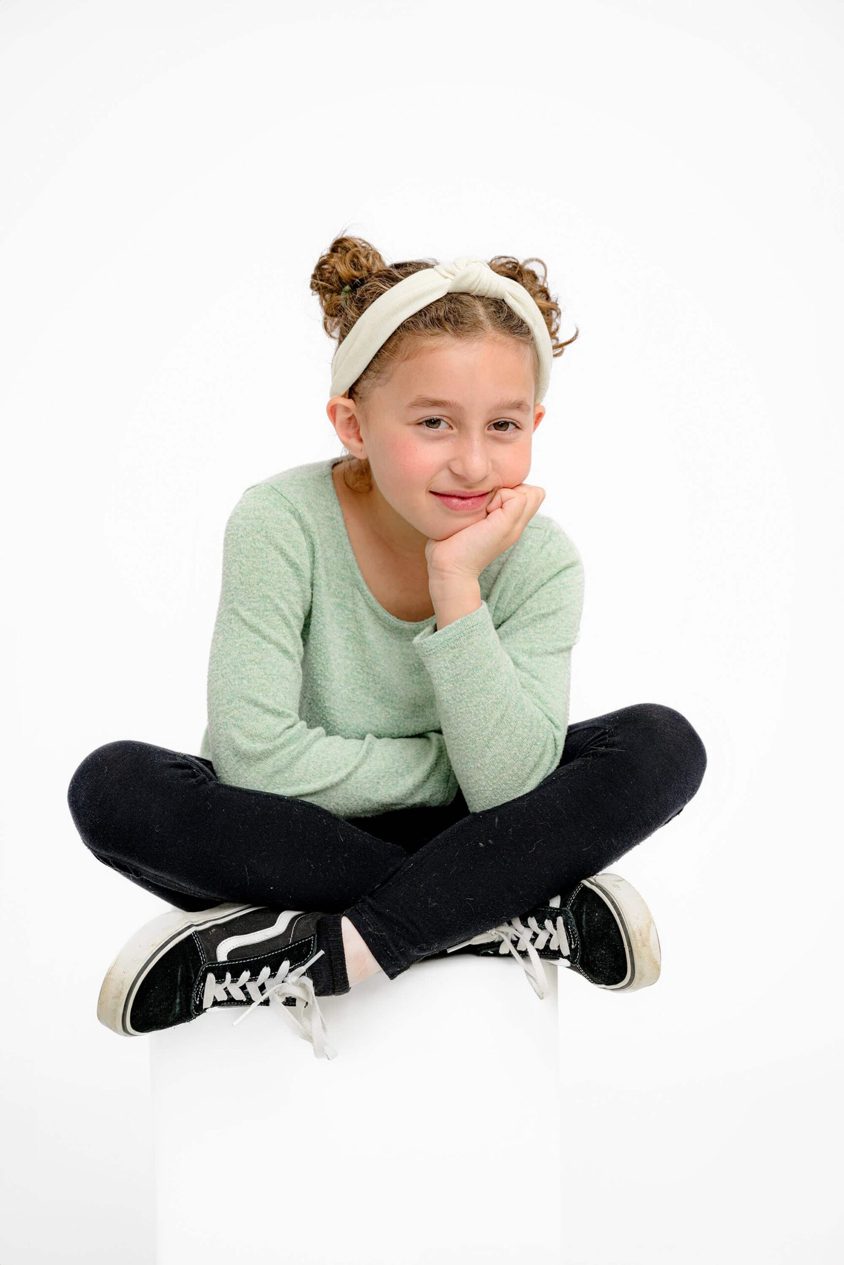 girl sitting cross legged on a stool with her hand on her cheek wearing a green sweater and black pants and shoes