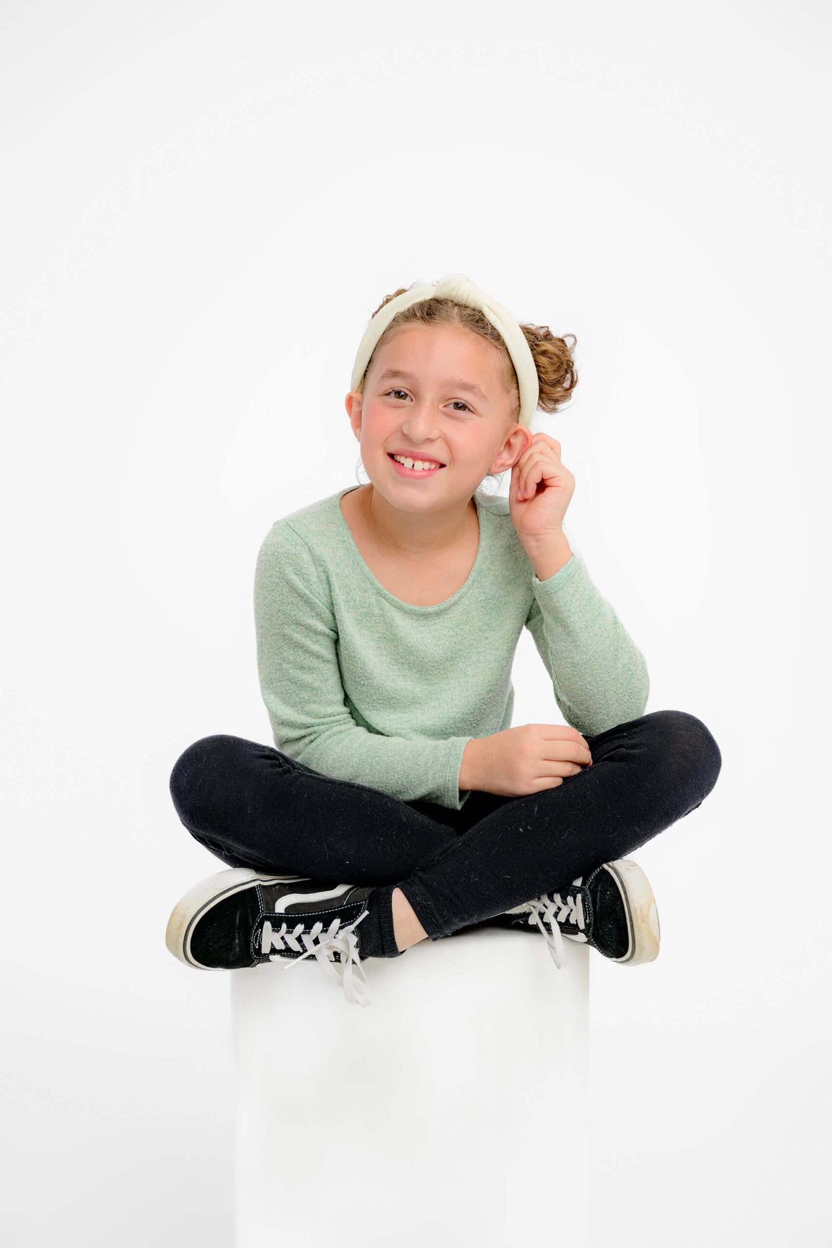 girl wearing a green shirt and black pants and shoes sitting cross legged tugging her ear