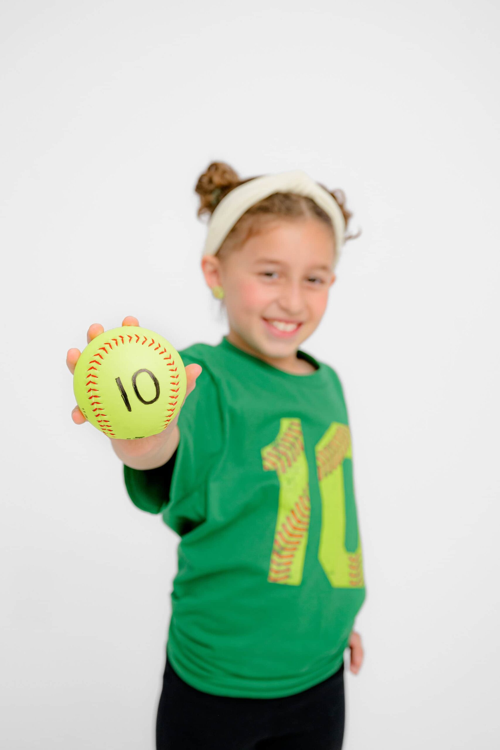 girl holds out a big yellow softball with the number 10 written on it to celebrate her upcoming birthday