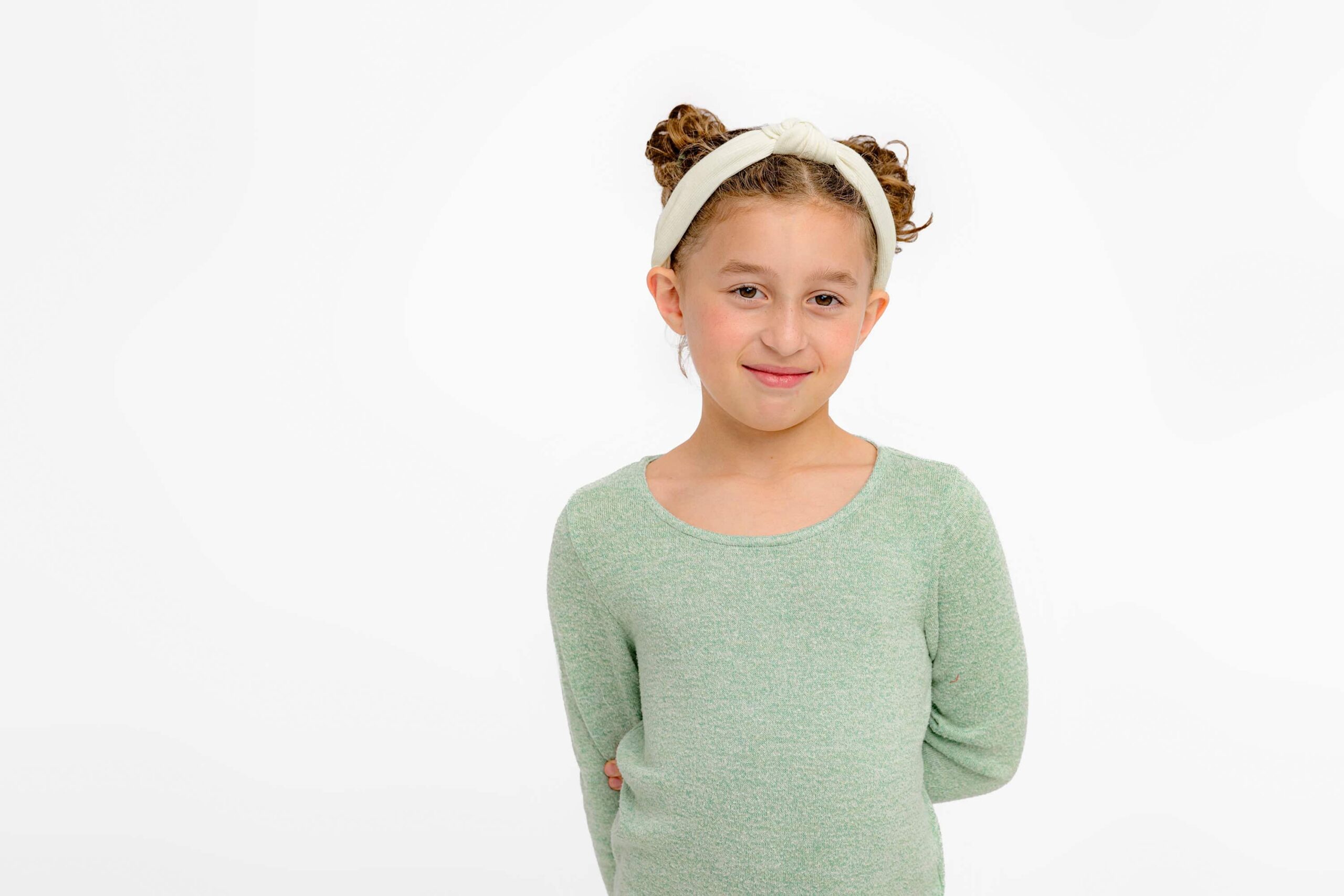 girl standing wearing a green shirt and headband with one arm tucked behind her back smiling gently