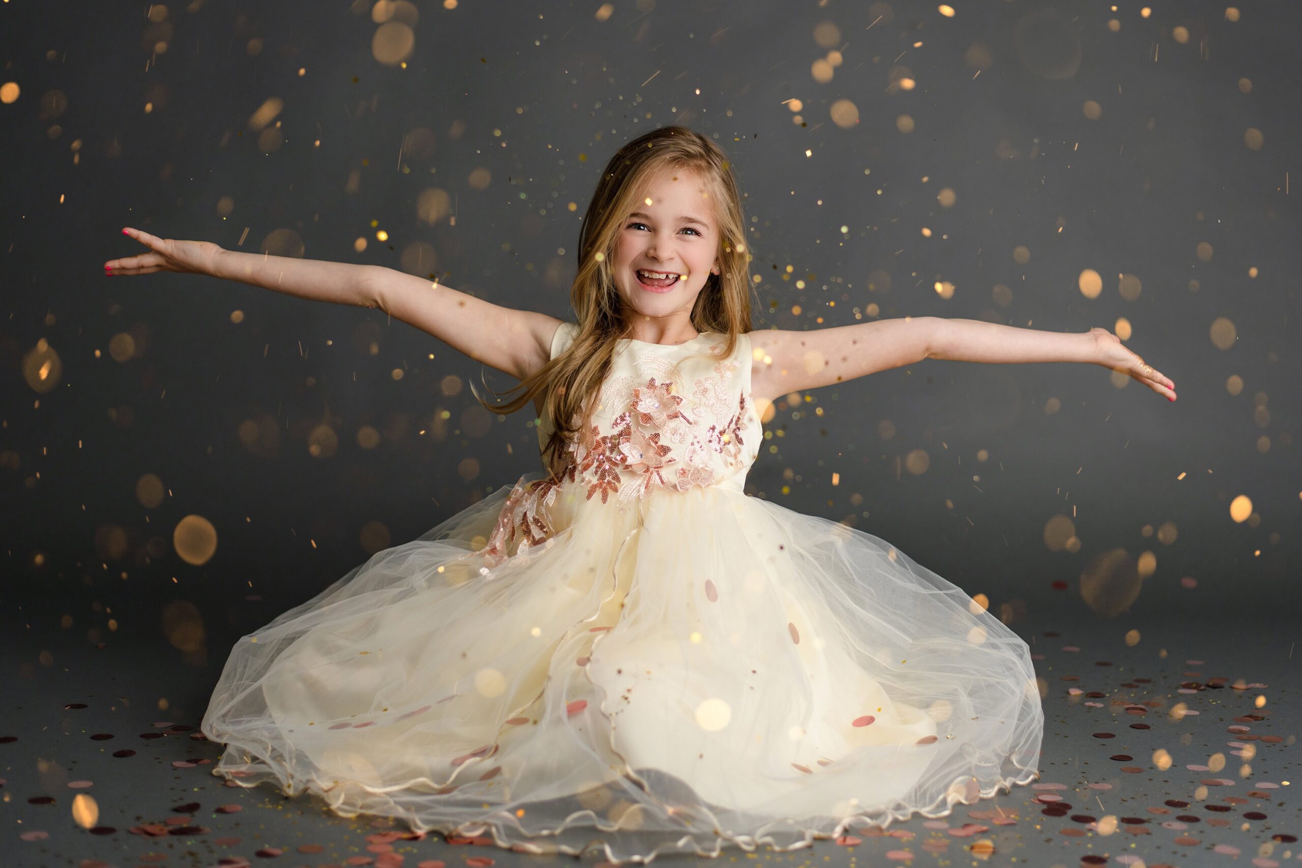 Young girl, dressed in cream with pink trim on a gray background, tosses gold glitter into the air with her arms open wide celebrating during her glitter mini session in the studio