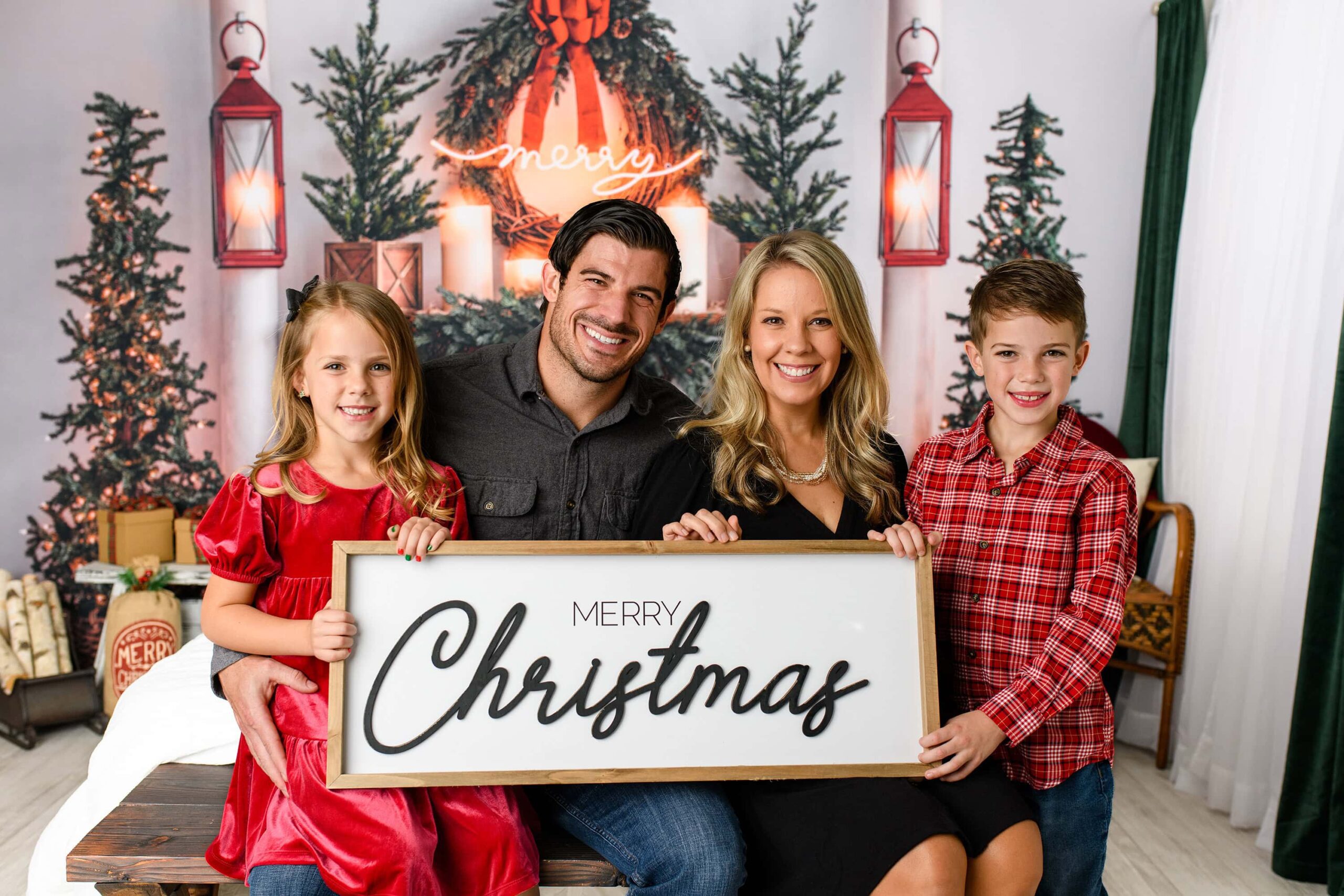 Family of four dressed in holiday colors hold a merry Christmas sign in their holiday, inspired photography session in a Waxhaw studio