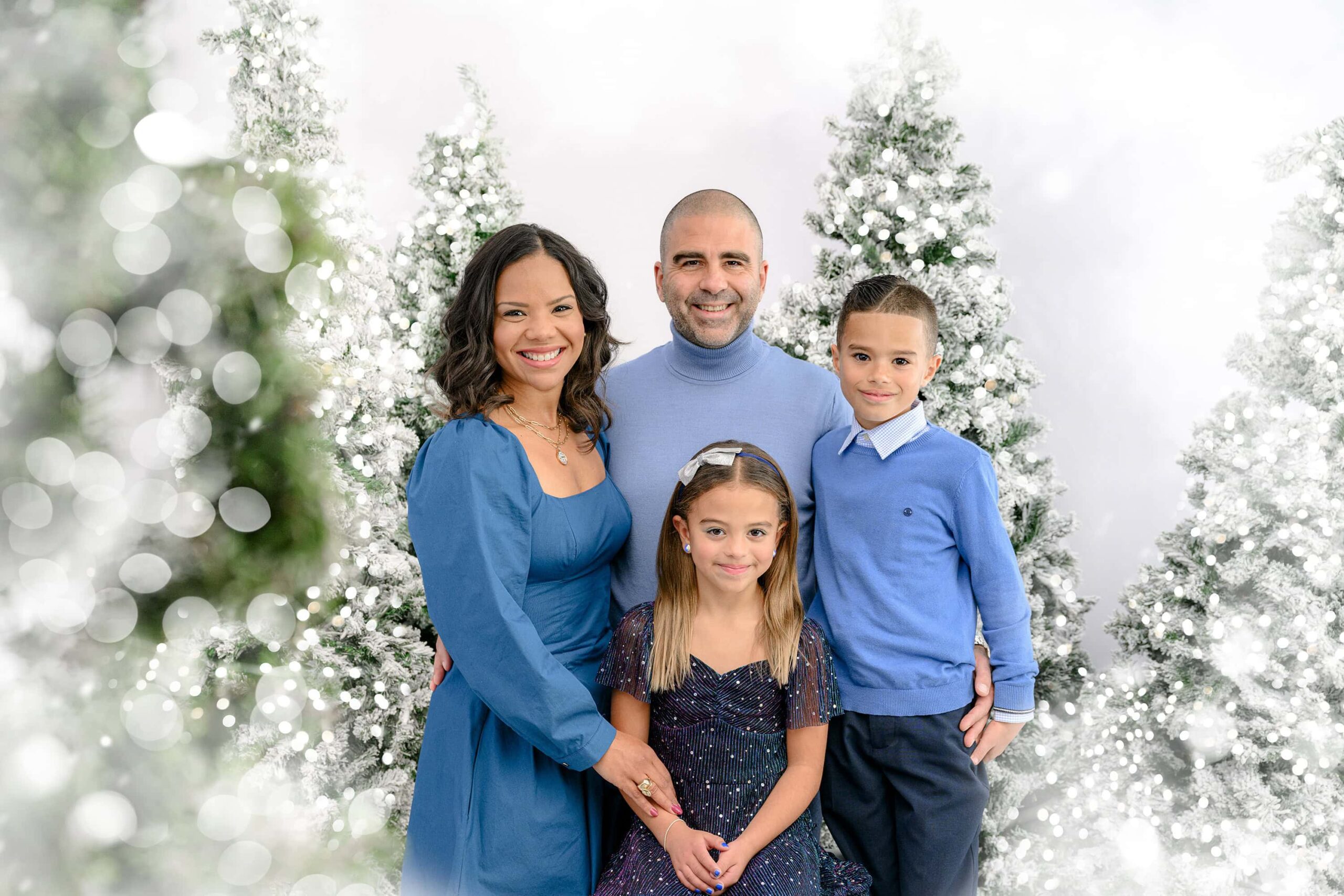 This family of four is dressed in blue tones surrounded by white Christmas holiday session set up in the Waxhaw North Carolina studio With white flocked trees and lights shining all around them