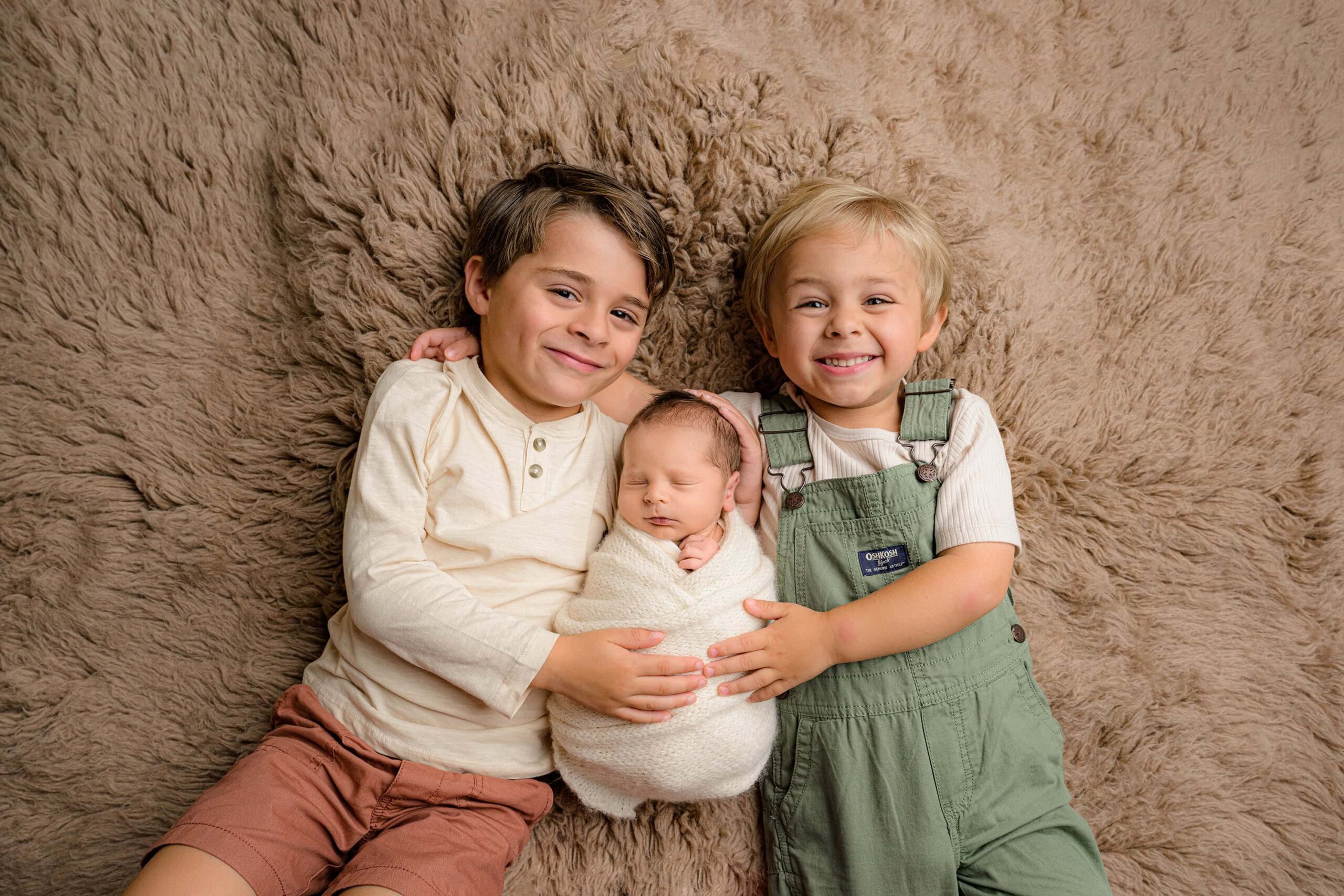 Three brothers dressed in similar warm tones lay together, celebrating their newborn session and welcoming the newest Baby brother and My Waxhaw, North Carolina photographystudio