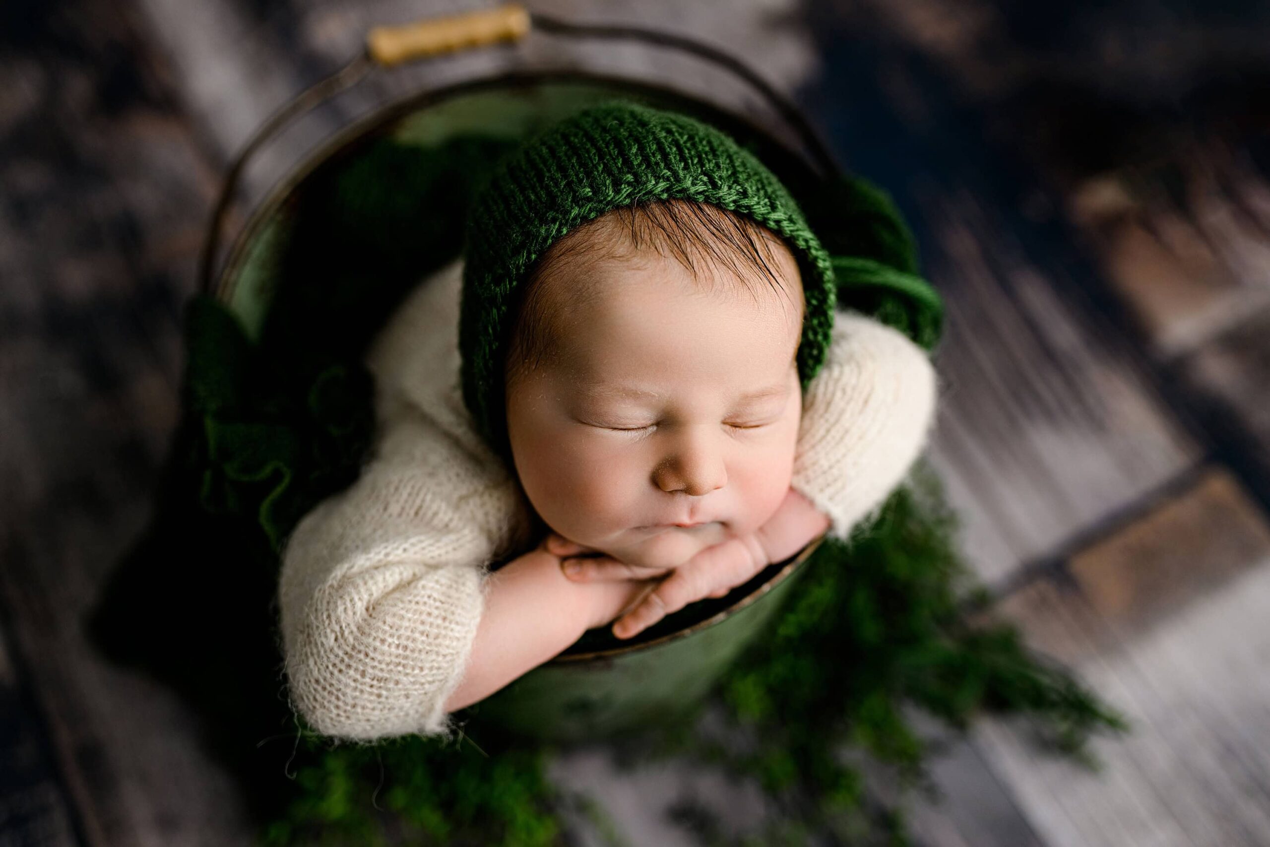 In a Waxhaw photography studio a Newborn boy is posed safely in a green bucket with his two hands under his chin wearing a green bonnet