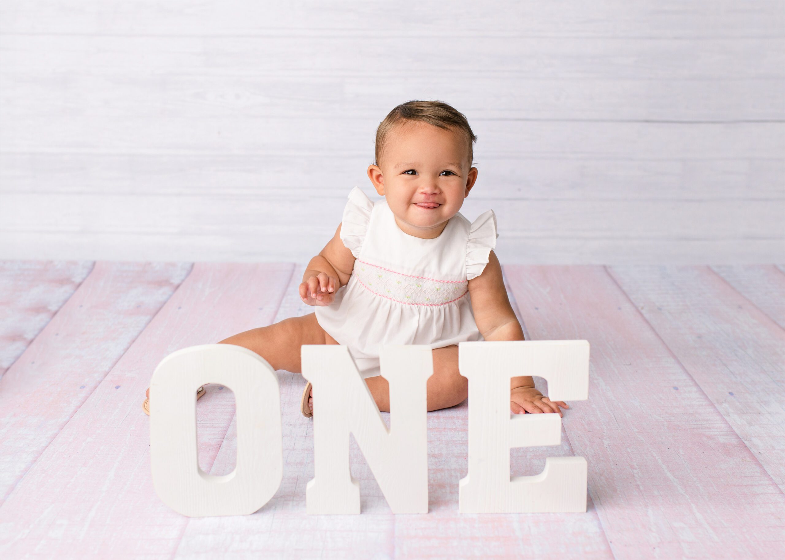 Pretty and pink this little girl dressed in white with a pink background sits during her one year-old milestone session With the letters, ONE in front of her