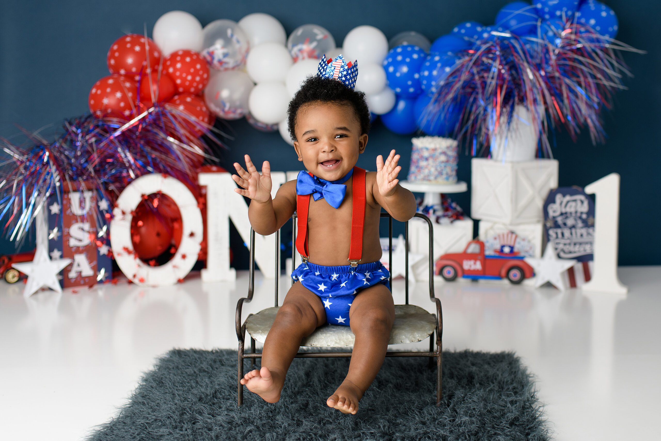 Little boy from Indian land, SC Proudly celebrates his Fourth of July birthday With a customized cake session in my Waxhaw, NC studio. He is wearing suspenders, a bowtie, and a diaper cover all in festive Fourth of July colors.