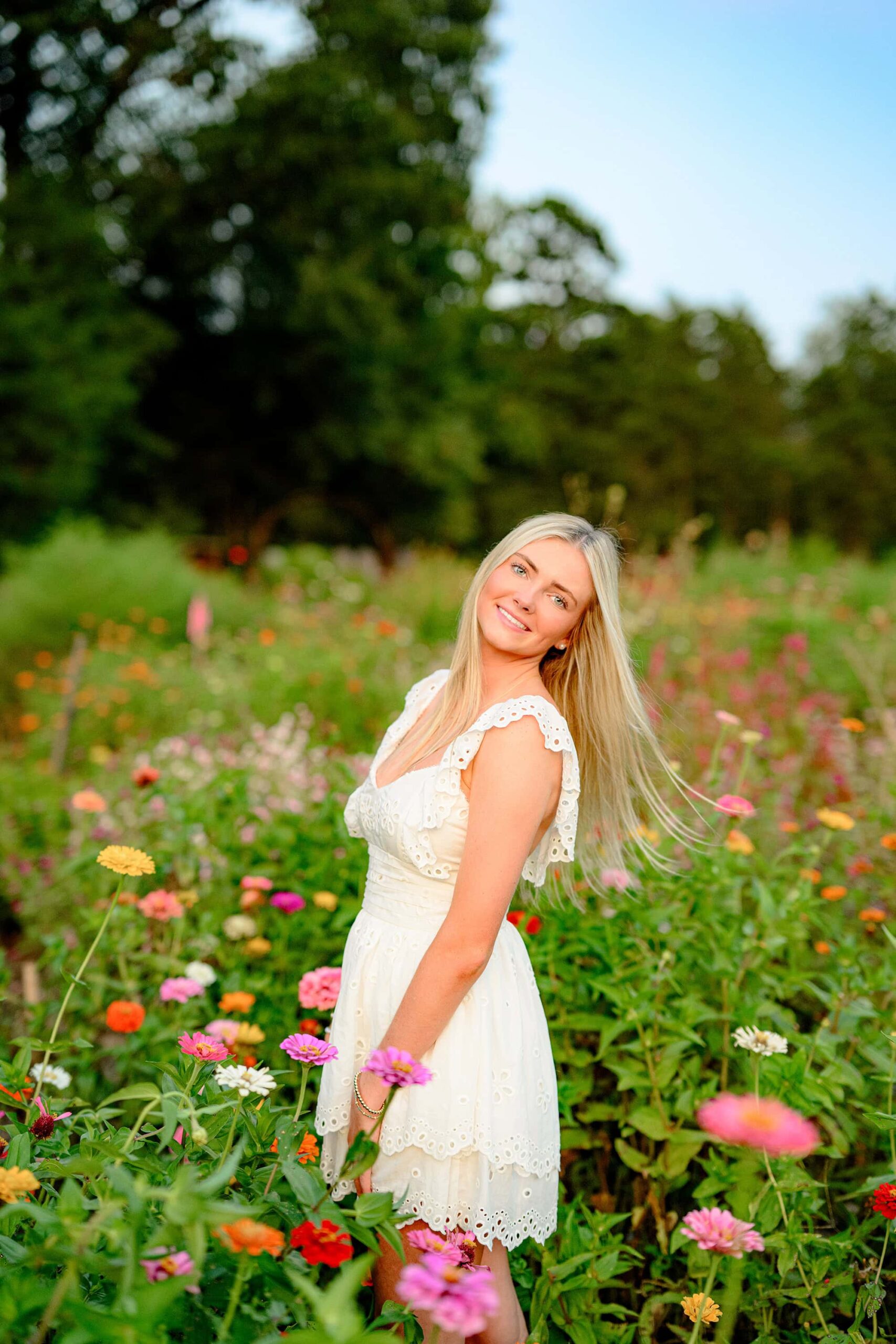A Cuthbertson senior from Waxhaw, NC, wearing a cream baby doll dress, twirls gracefully in a bright and colorful flower field in Lancaster, SC. Her hair flows in the wind, creating a dynamic and joyful moment.