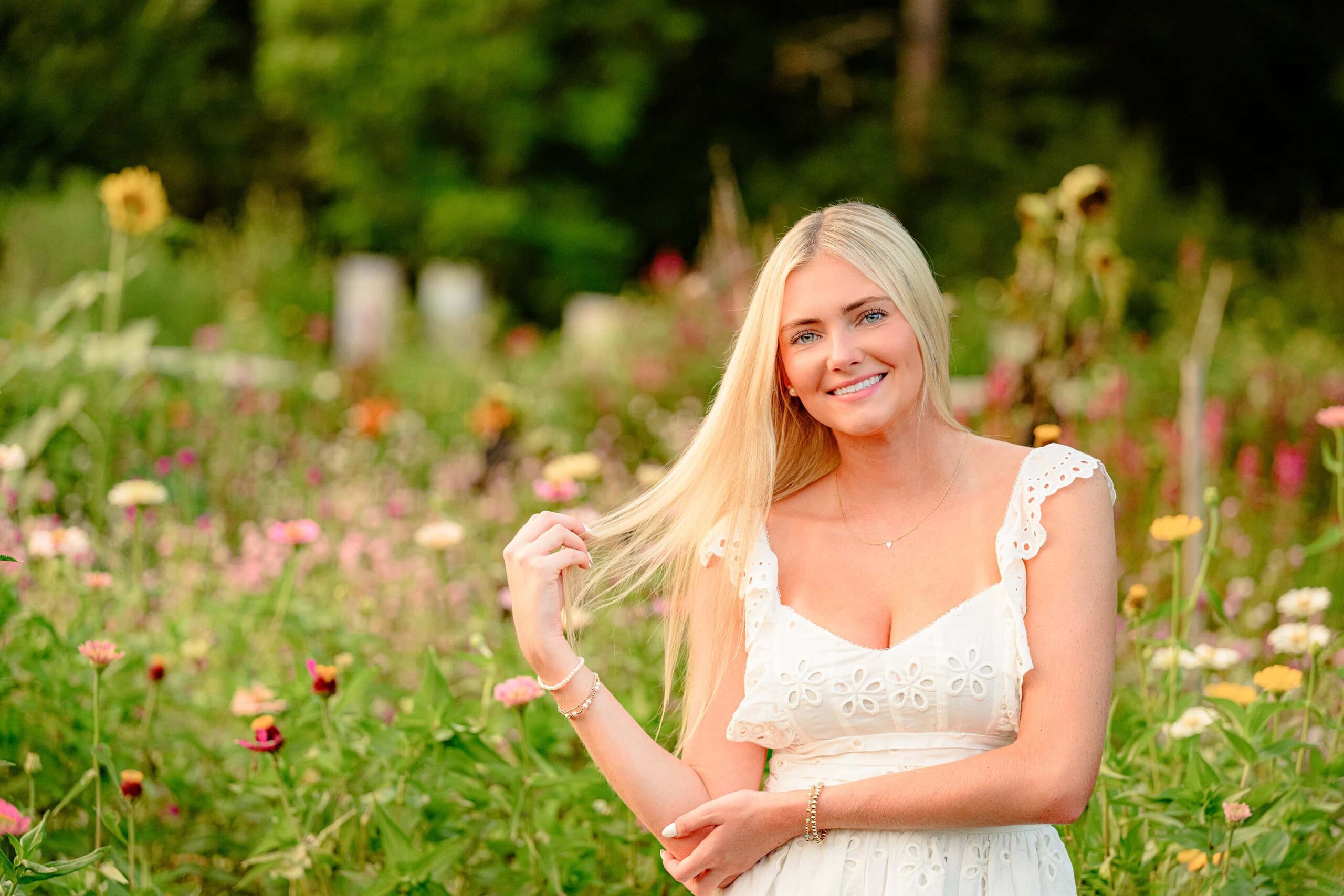 A vibrant and picturesque flower field in full bloom, bursting with an array of bright colors under a clear blue sky. The flowers stretch endlessly, creating a dreamy backdrop for photography sessions. In the foreground, a professional photographer engages with a senior wearing a cream baby doll dress, capturing natural poses like twirling, walking, and playful gestures. The golden hour sunlight casts a warm, enchanting glow, adding magic to the moment. This scene highlights the perfect timing of the session, capturing the peak bloom season of the field. Book a session now to experience the beauty of Lancaster, SC's flower fields and create timeless memories with an experienced photographer who helps bring your vision to life.
