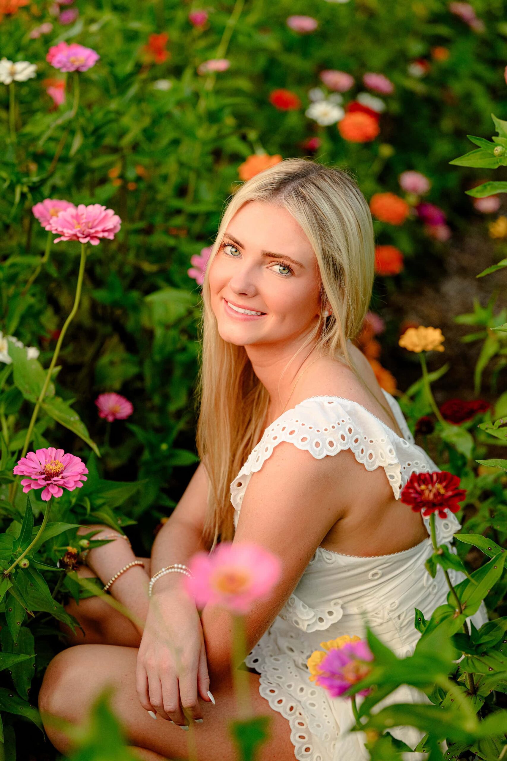 The senior is squatting down, surrounded by colorful flowers. Her dress gently spreads around her, and her relaxed pose blends beautifully with the vibrant blooms.