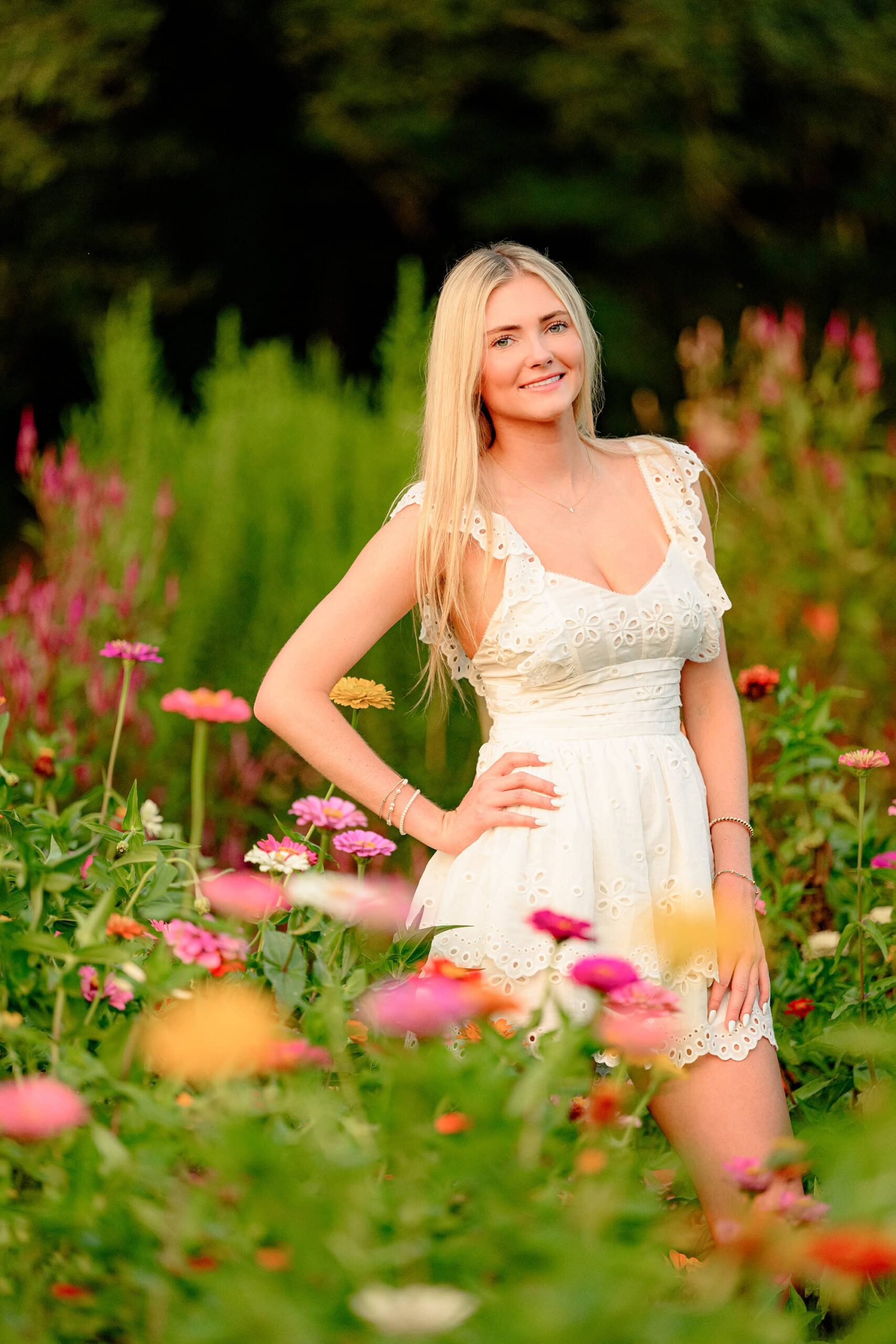 The senior stands confidently in the flower field with one hand on her hip. Her cream baby doll dress complements the bright and lively colors of the blooms around her.