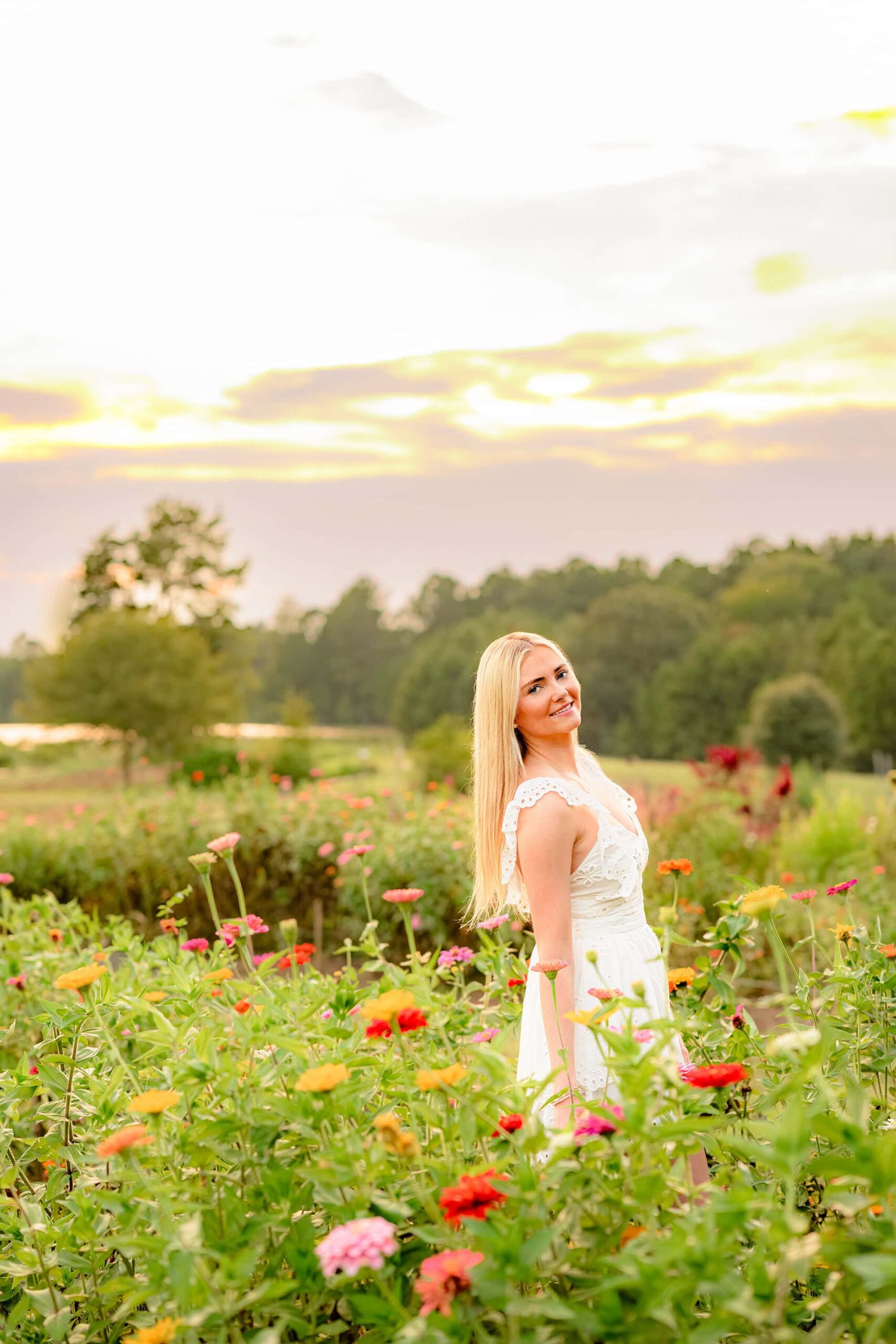 The senior stands in the vibrant flower field with a stunning sunset glowing behind her, casting a golden light on her cream baby doll dress and highlighting her serene expression.