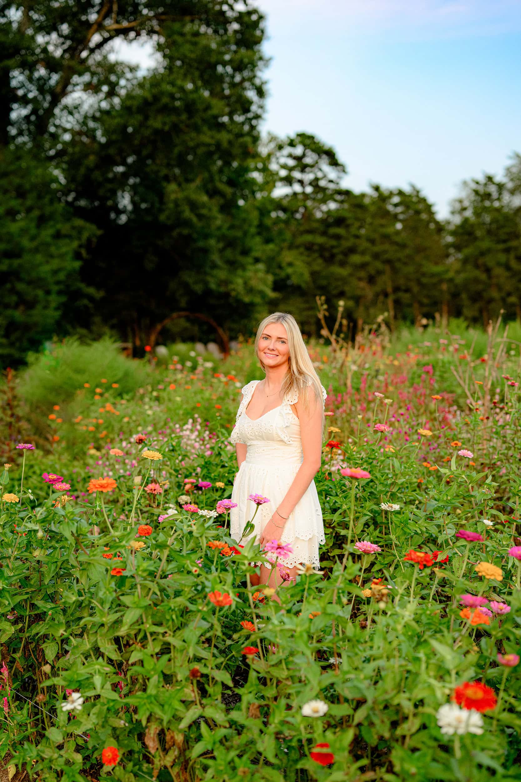 The senior is fully immersed in the colorful flower field, standing amidst the blooms with her dress gently flowing, creating a harmonious and picturesque scene.