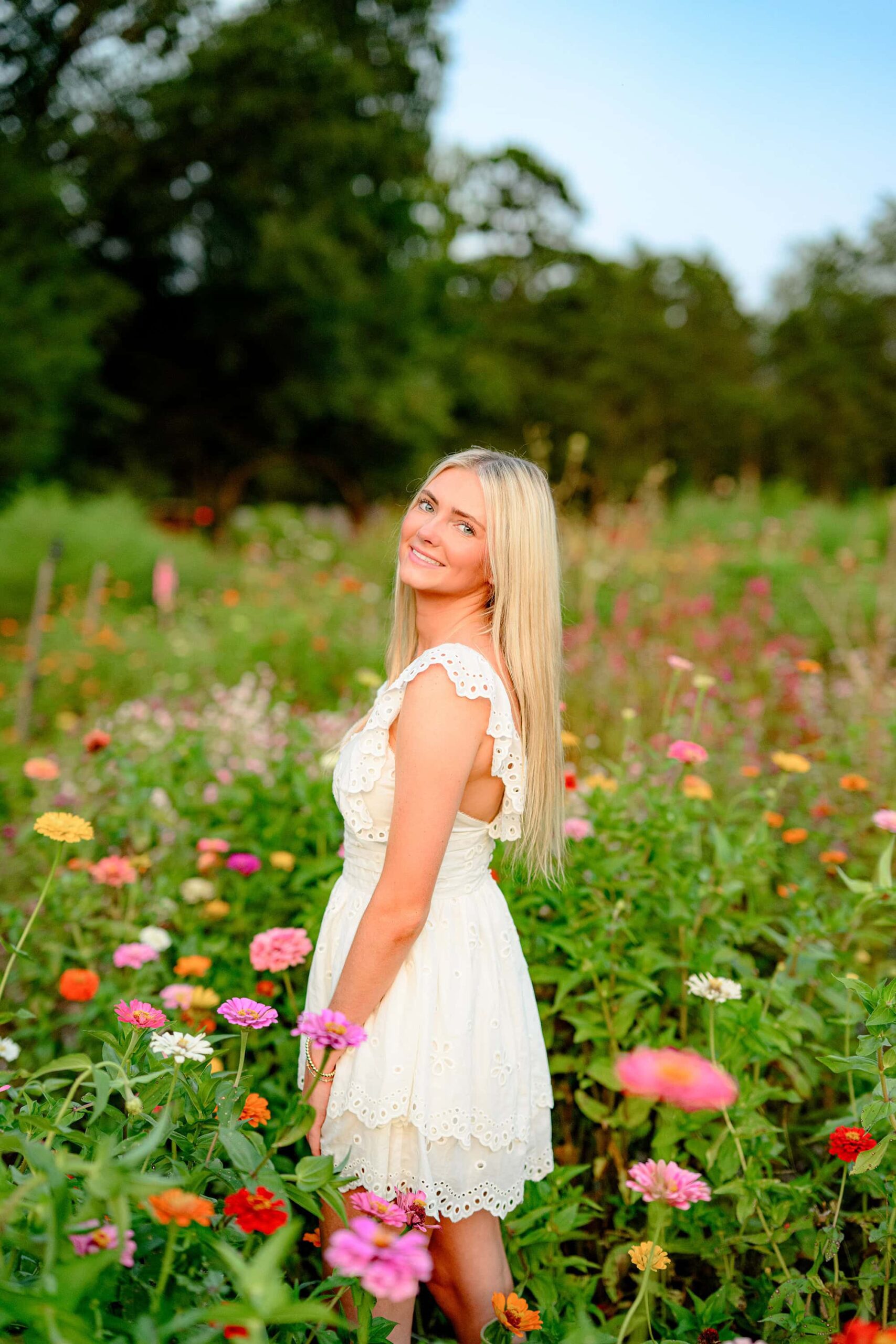 The senior glances over her shoulder with a soft smile, her cream baby doll dress catching the light as the vibrant flowers create a colorful and dreamy background.