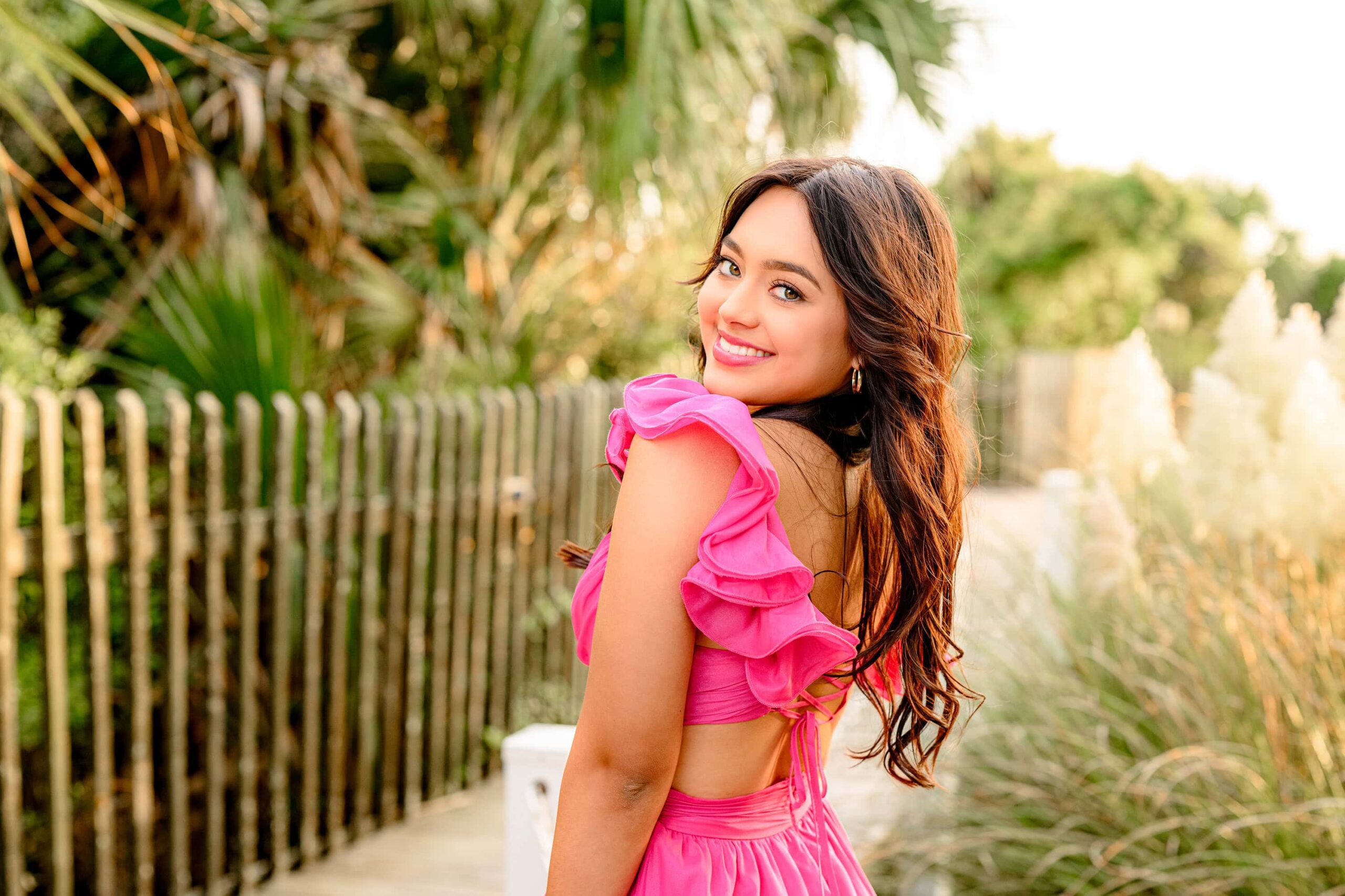 A senior with long dark hair and bright green eyes, smiling as she looks over her shoulder while wearing a pink dress with flutter sleeves and a detailed back, with a scenic boardwalk and ocean in the background.