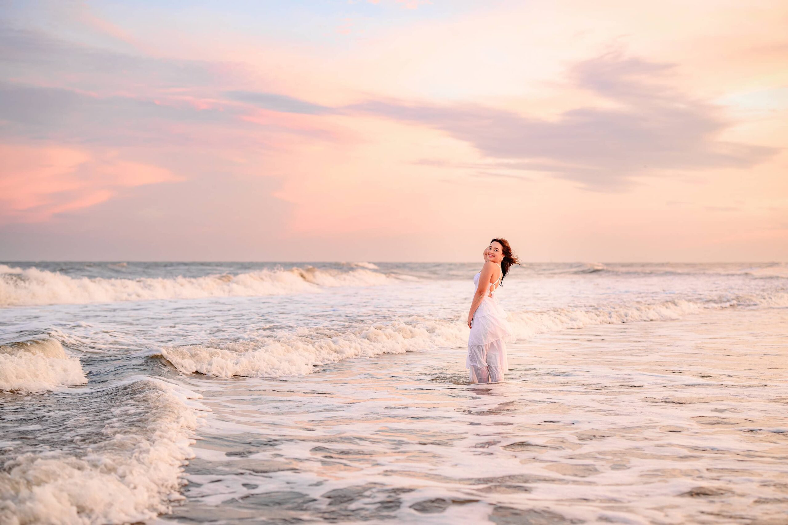 A Cuthbertson High School grad, with long dark hair and bright green eyes, standing in the ocean with waves crashing around her, her hair blowing in the wind, wearing a flowing white dress for a serene and dramatic beach moment.