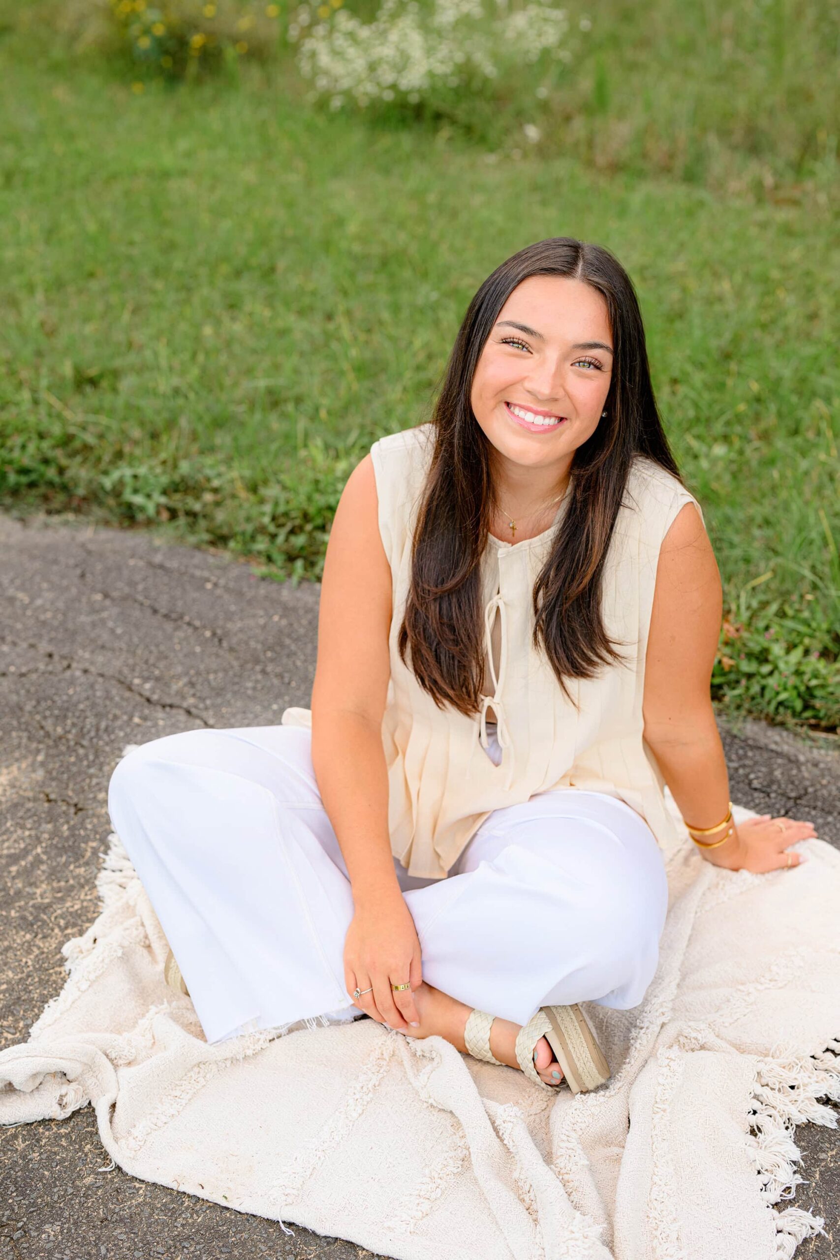 A relaxed and cheerful shot of the senior girl in white pants and a cream top, sitting on a blanket in the park and smiling at the camera, radiating warmth and joy.