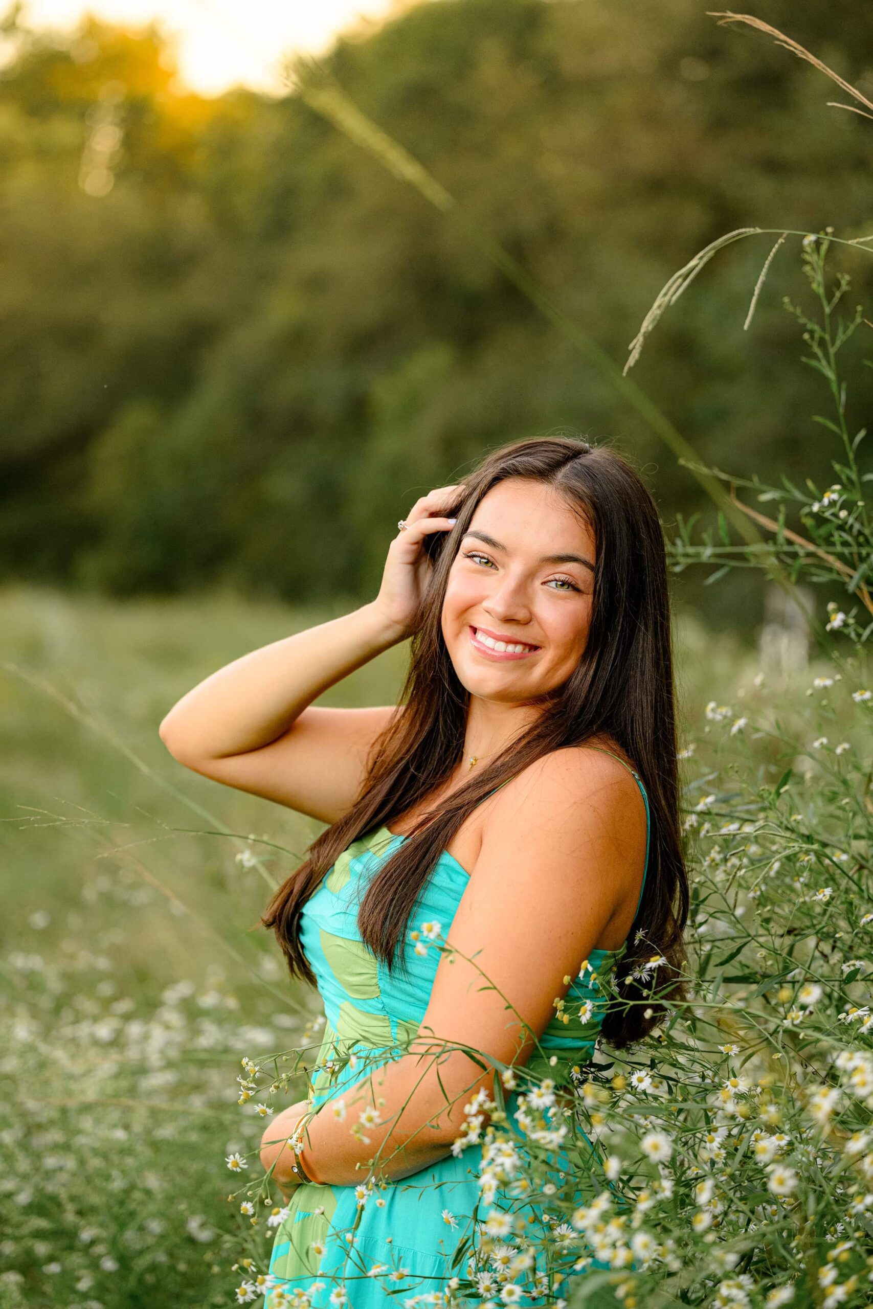 A senior girl with long dark hair, wearing a teal dress, gently touching her hair while standing in a park surrounded by vibrant flowers, creating a serene and elegant moment.