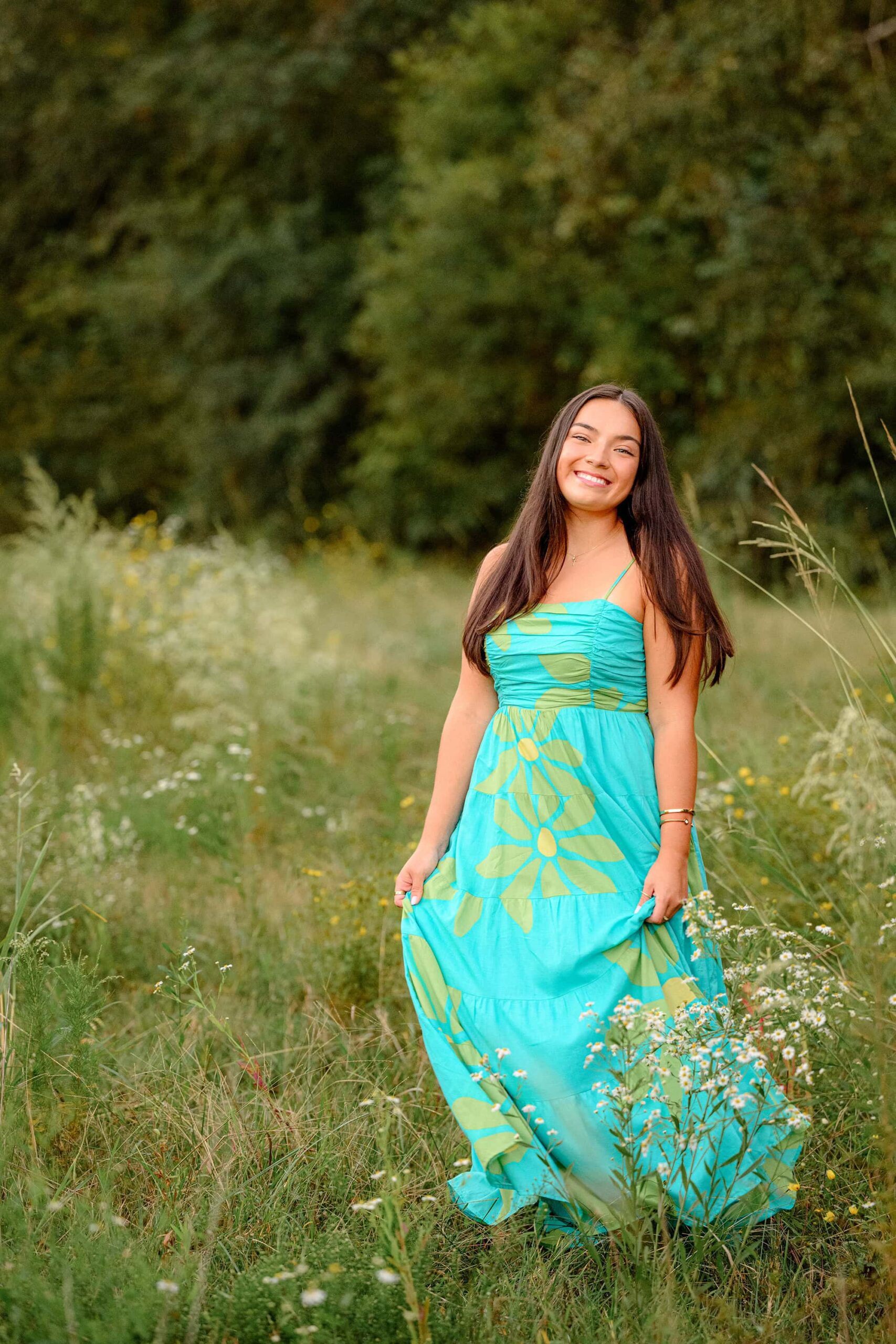 The same senior girl in a teal dress with small straps, playfully adjusting her dress while standing in tall grasses, captured in a moment of carefree beauty and natural surroundings.