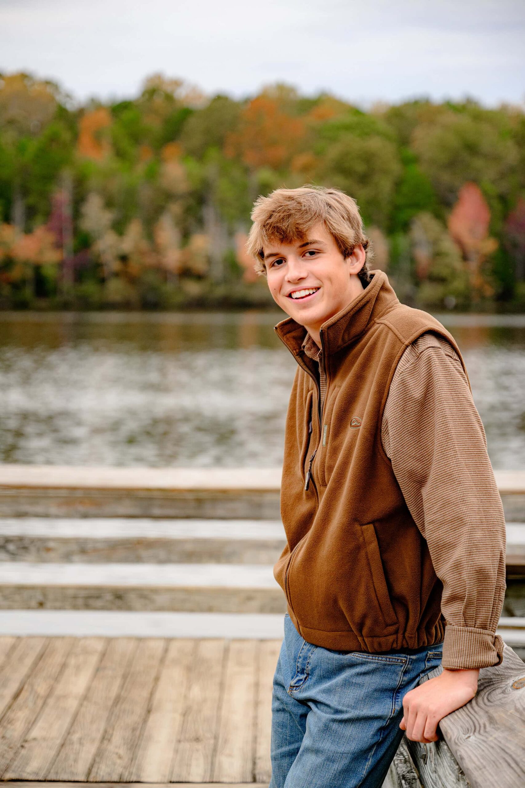 A senior boy from Lancaster, SC, looking directly at the camera while leaning against a wooden rail, with vibrant fall trees and a tranquil pond in the background, exuding a relaxed and confident vibe.