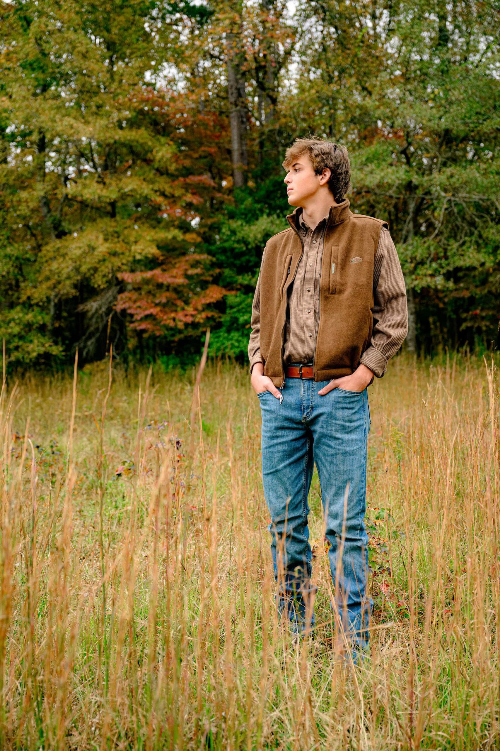 A senior boy from Lancaster, SC, dressed in a brown vest and plaid shirt, gazing off to the side while standing amidst tall grasses, creating a thoughtful, serene moment.
